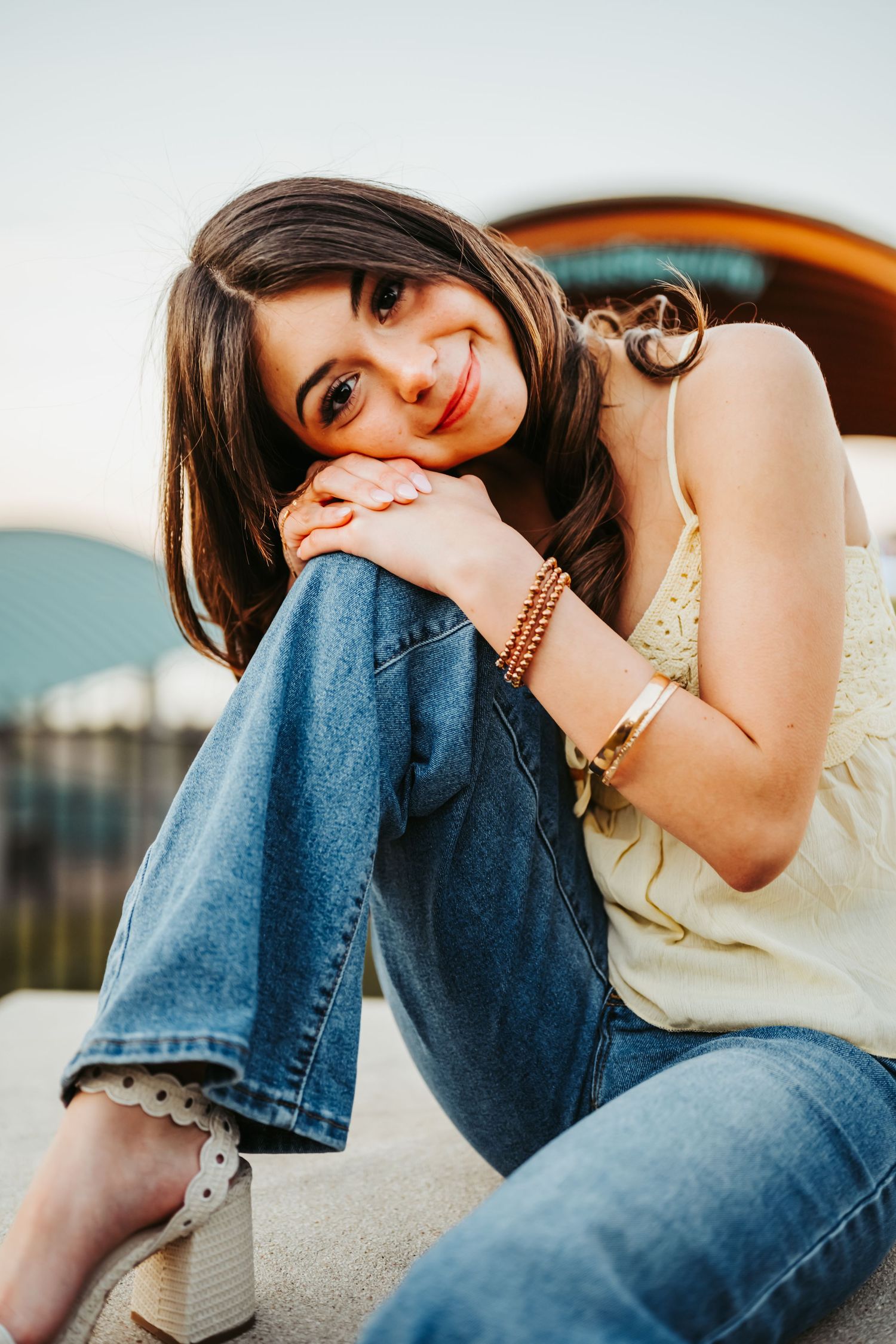 Young woman in jeans and yellow top sitting outdoors, smiling warmly at camera during golden hour.