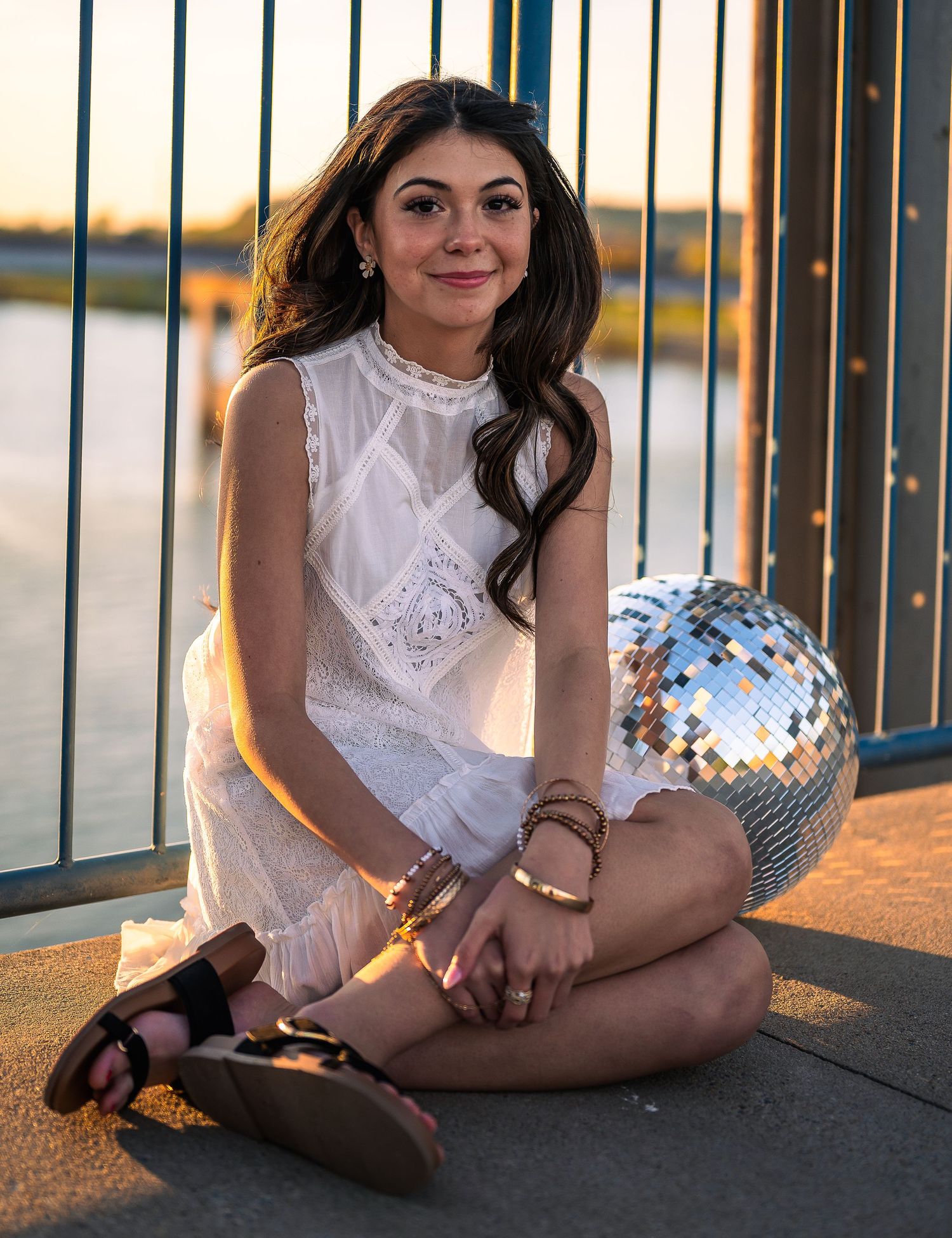 Smiling young woman in white sleeveless dress sitting cross-legged beside a disco ball at golden hour.