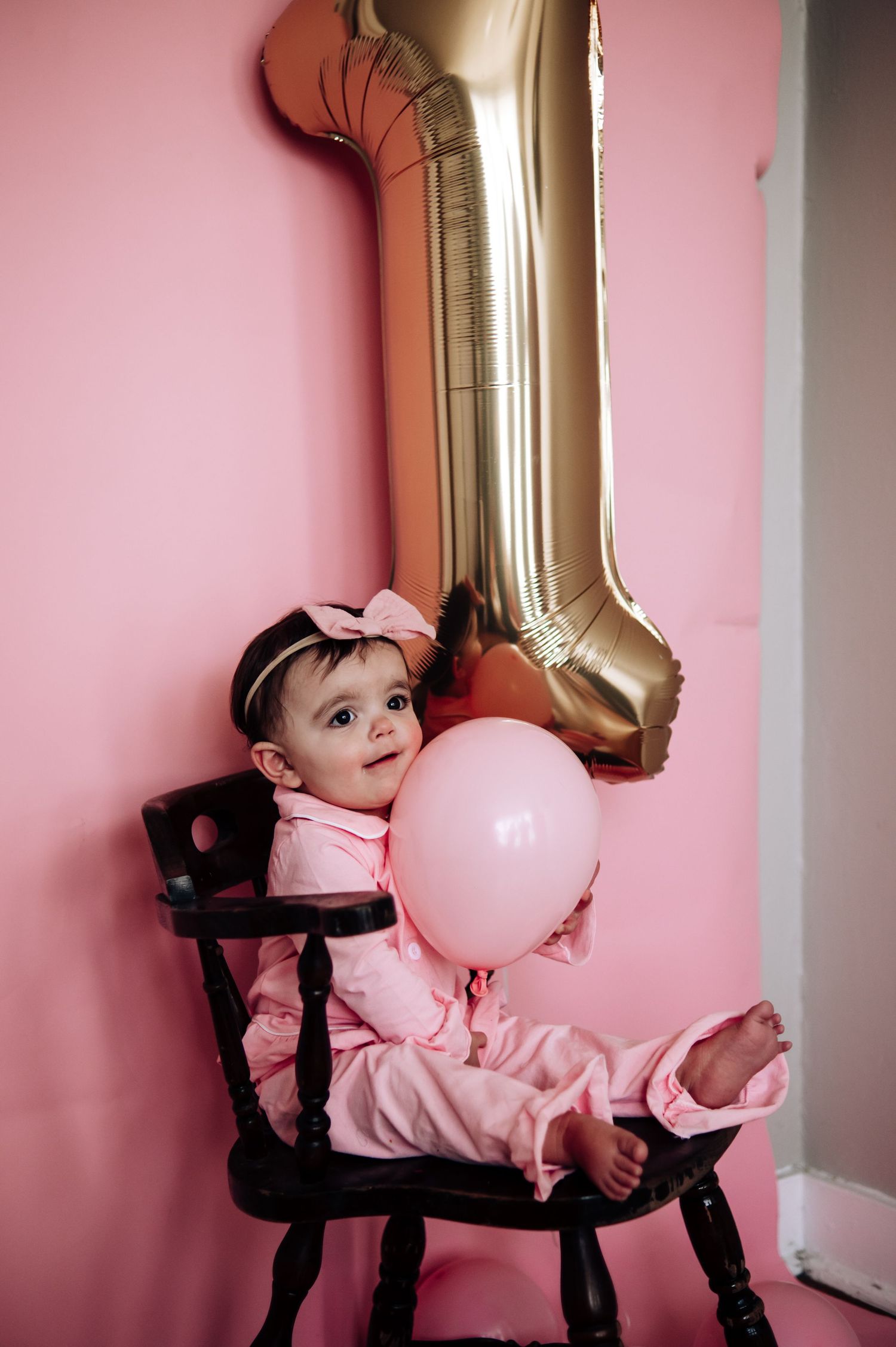 Baby in pink dress reclines on black chair with gold balloon and pink decor against rose-colored wall.