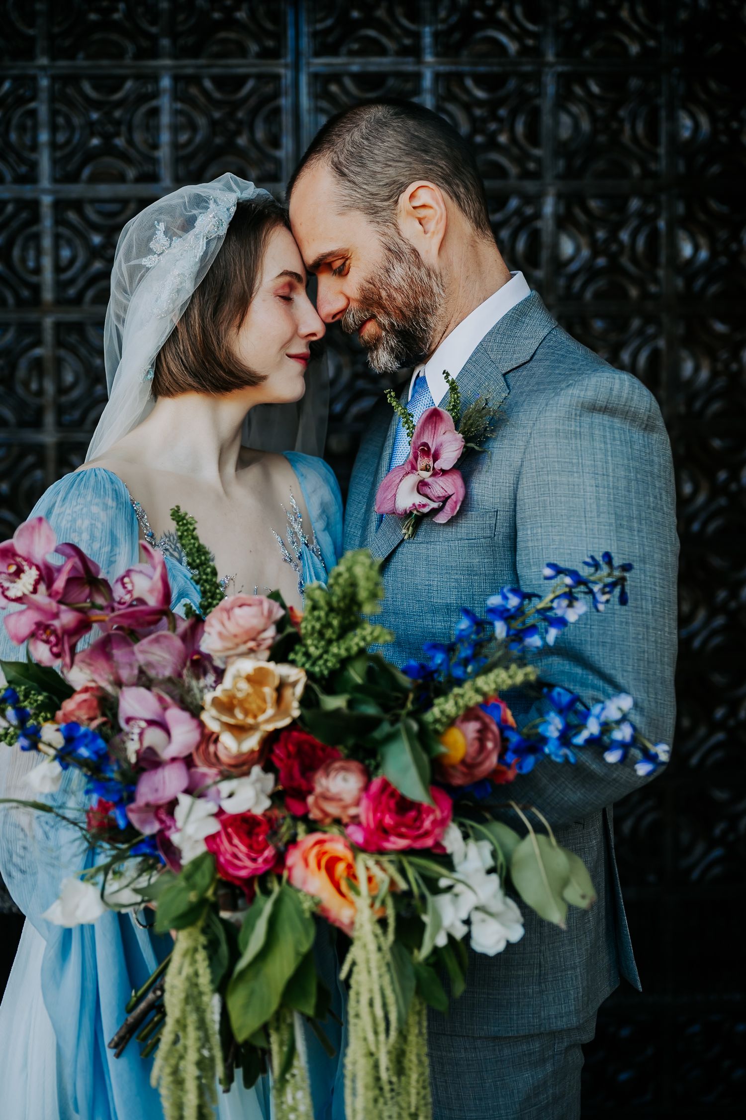 A romantic wedding couple poses with a vibrant bouquet featuring pink, blue and orange flowers against an ornate backdrop.