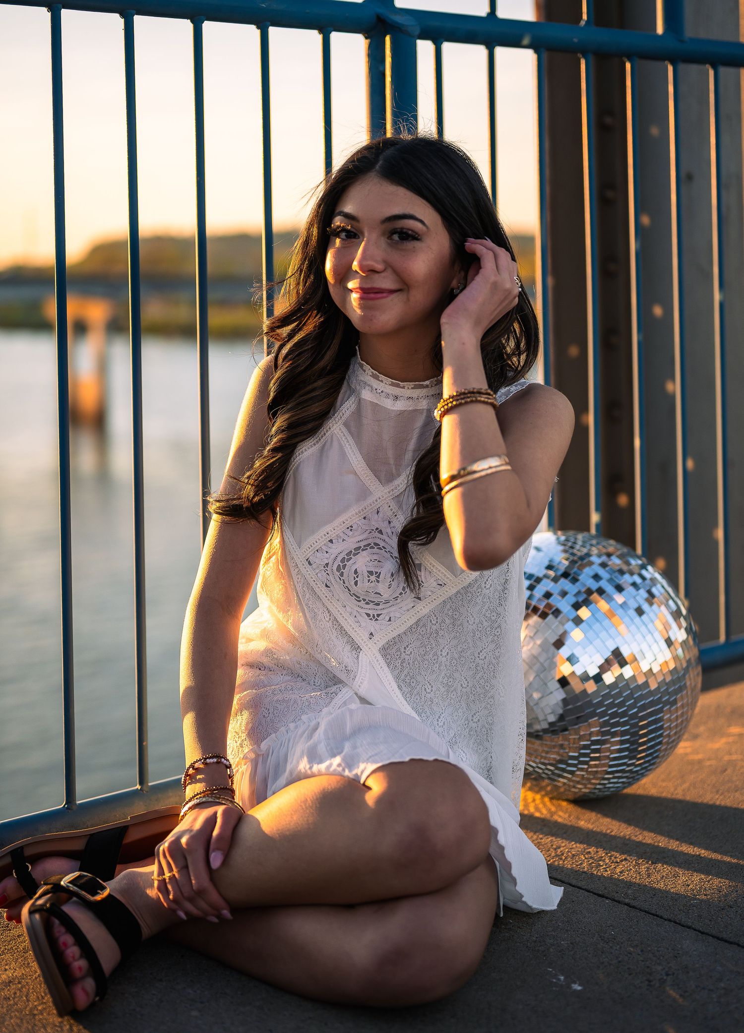 Young woman in white lace dress sitting by a railing at sunset with a disco ball nearby.