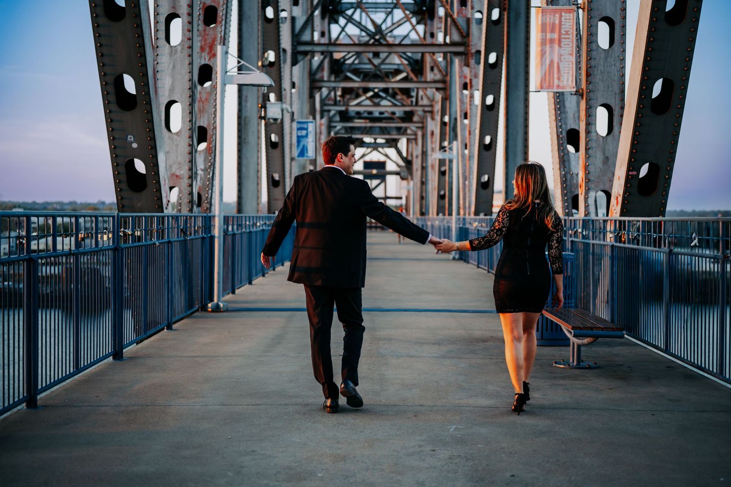 Couple holding hands walking on a steel bridge at dusk, dressed in elegant black attire.