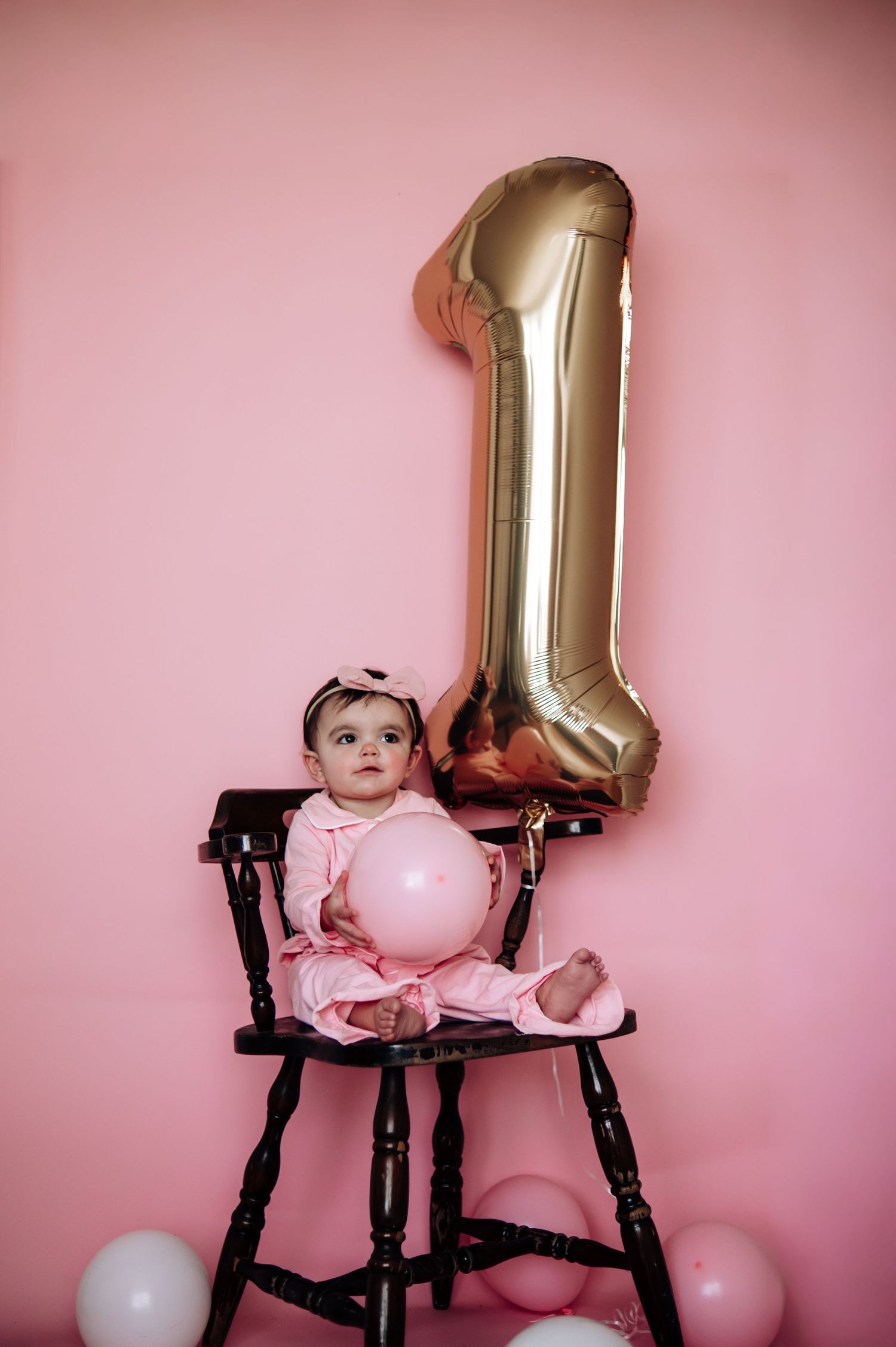 Baby in pink dress sits in antique high chair with gold number one balloon and pink decor for first birthday celebration.