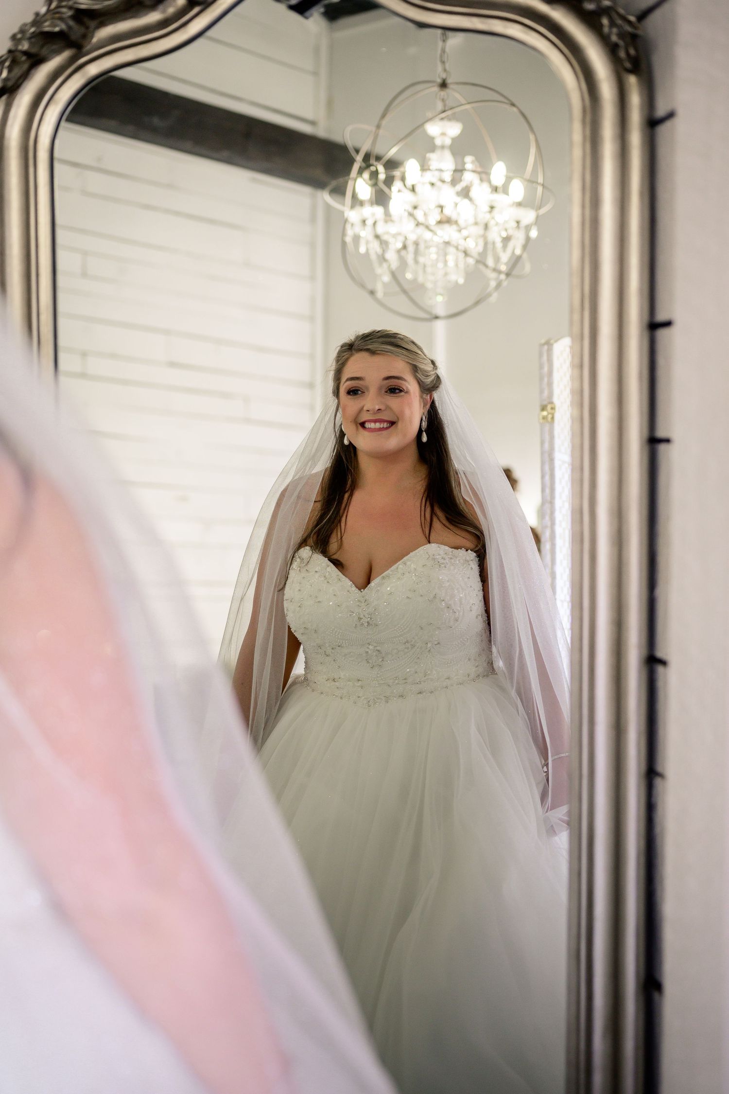 Bride in a strapless lace ballgown and veil smiles at her reflection in an ornate silver mirror with chandelier above.