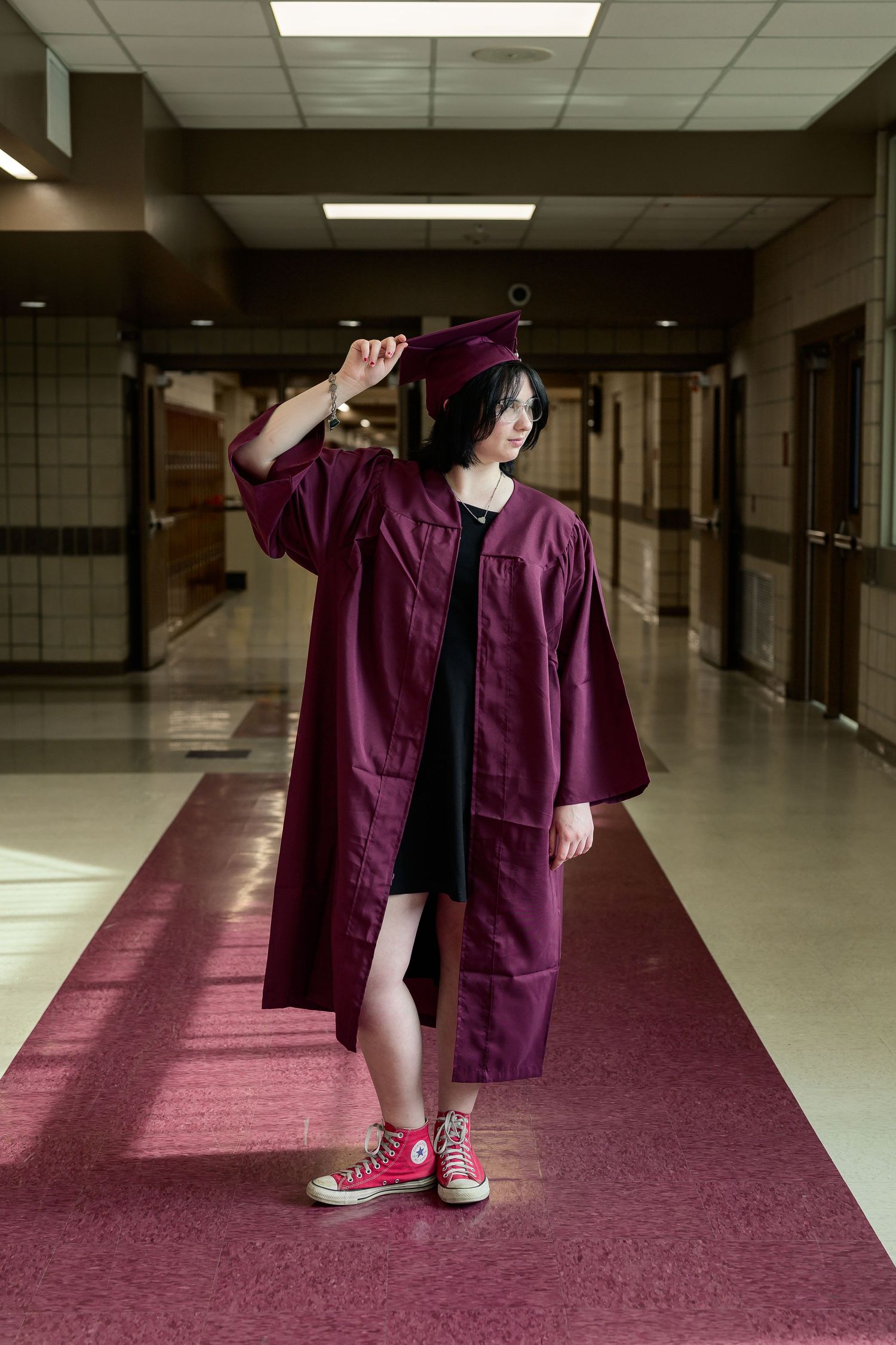 Graduate in maroon cap and gown posing in school hallway wearing red Converse sneakers.