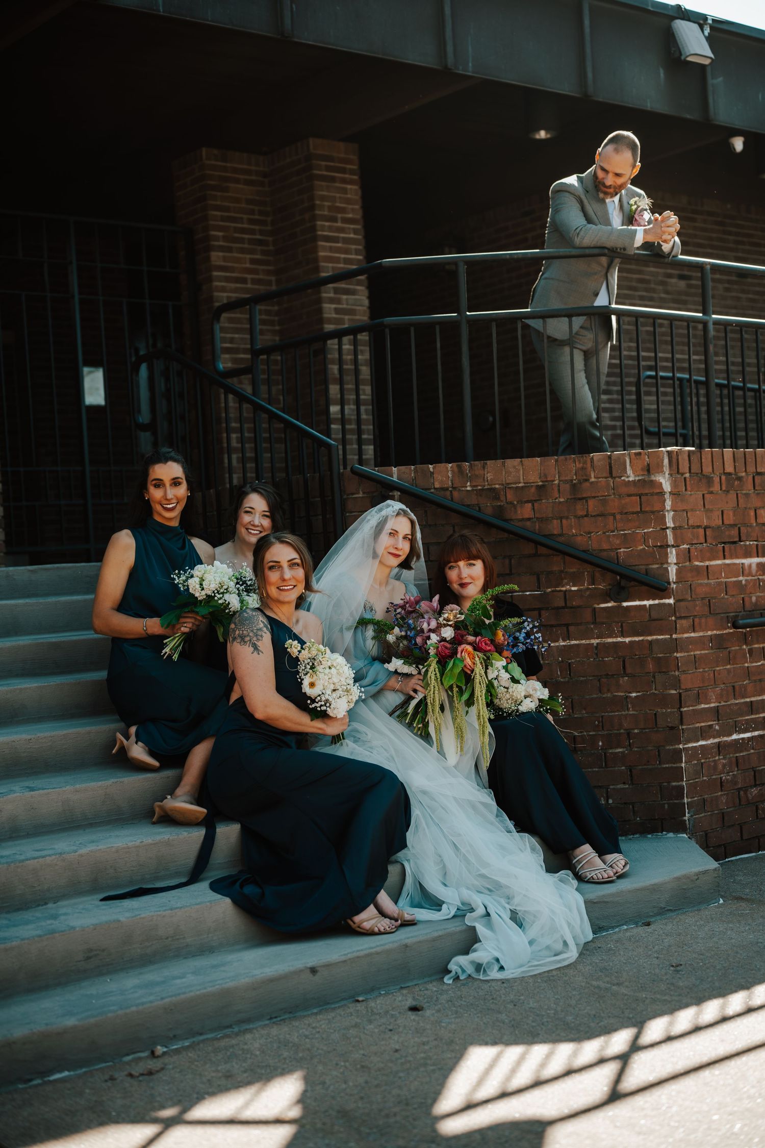 Wedding party sits casually on brick steps in natural light, sharing candid laughter and conversation.