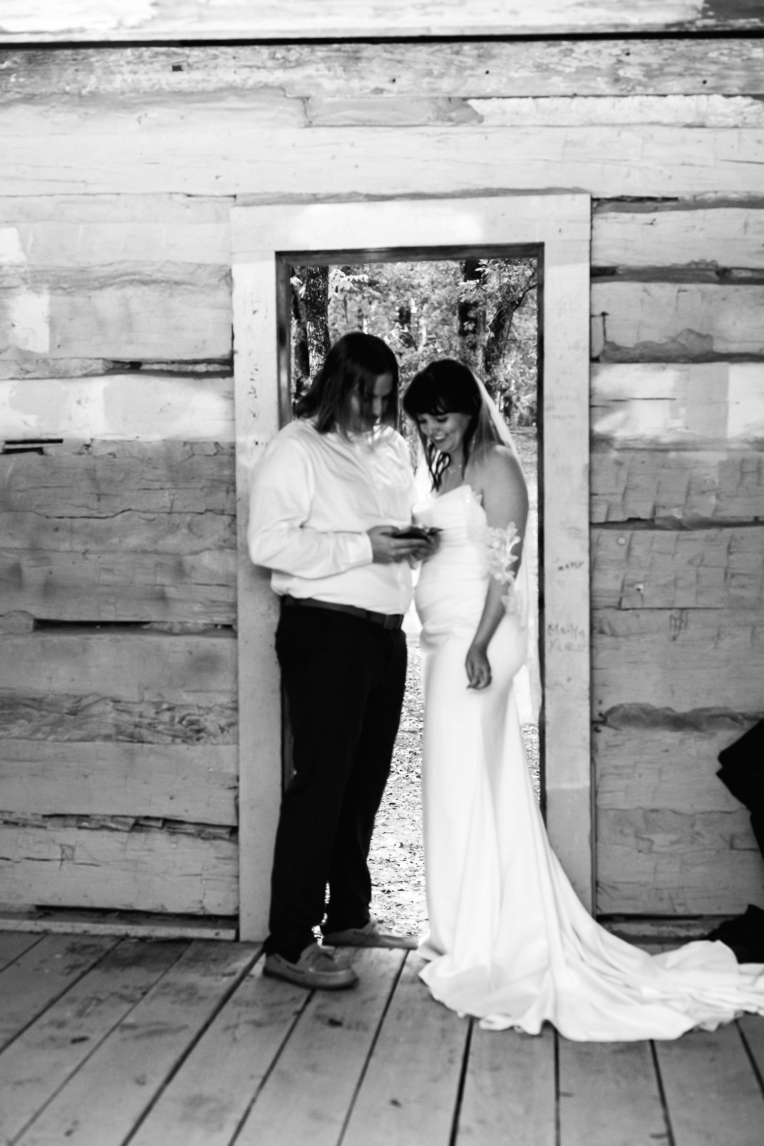 A black and white photograph showing two people in a rustic wooden doorway, one wearing a wedding dress with a long train.