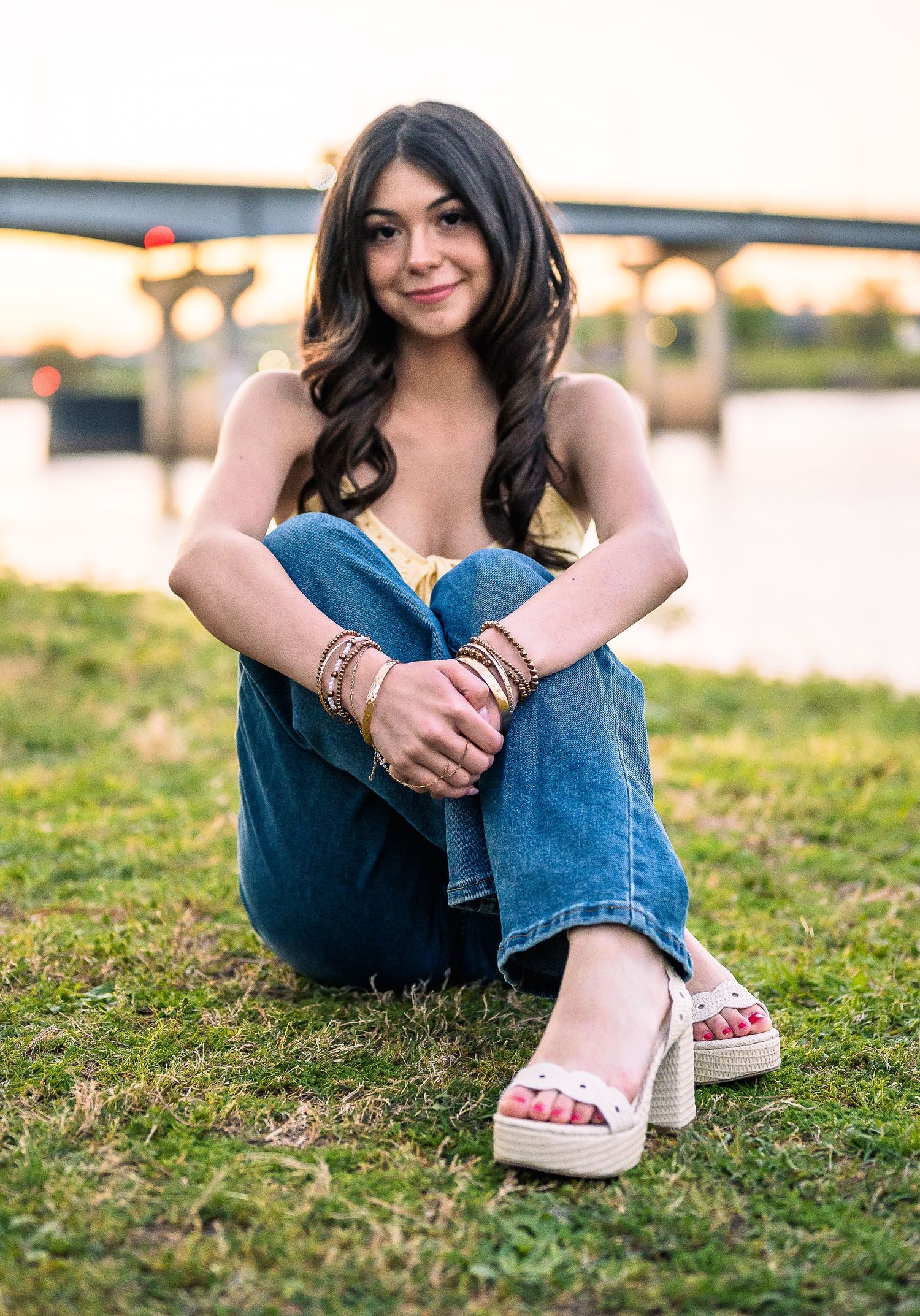 Young woman with dark hair sits on grass by a river at sunset, wearing jeans, yellow top, and white platform sandals.