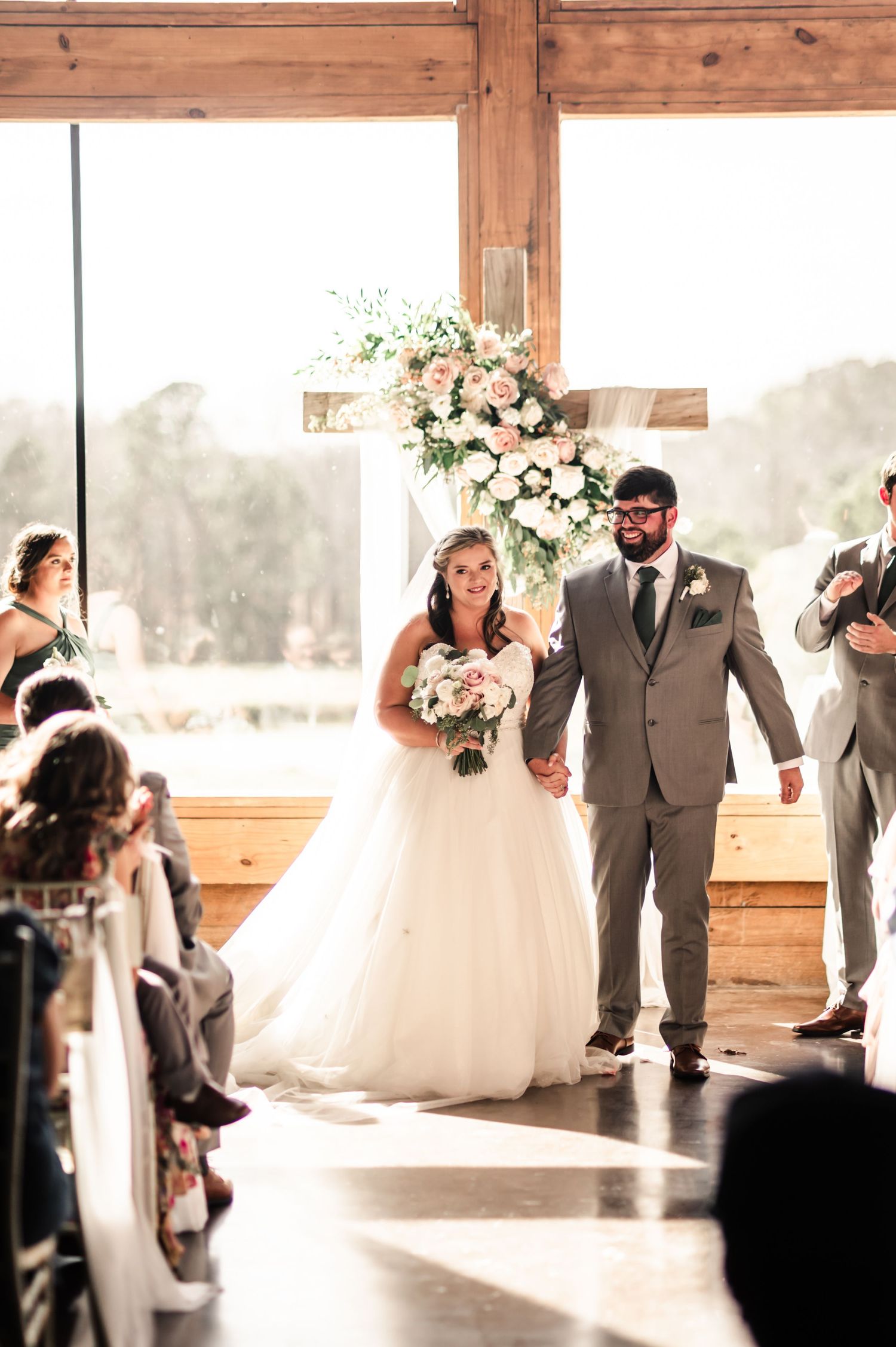 Bride and groom walk down the aisle at an indoor barn wedding ceremony with a floral cross backdrop.