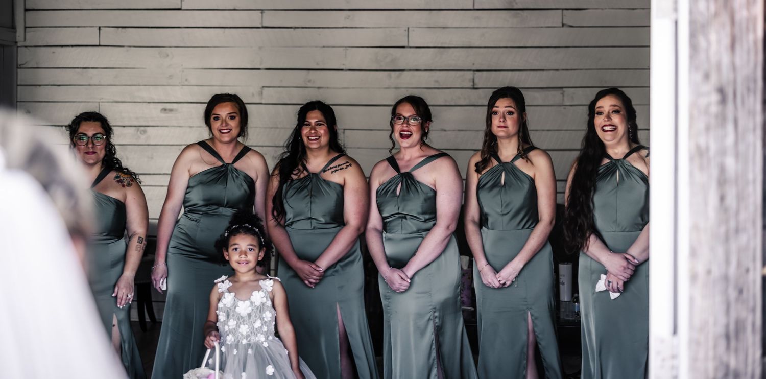 Bridesmaids in sage green gowns react emotionally as bride approaches, with flower girl in gray dress at wedding.