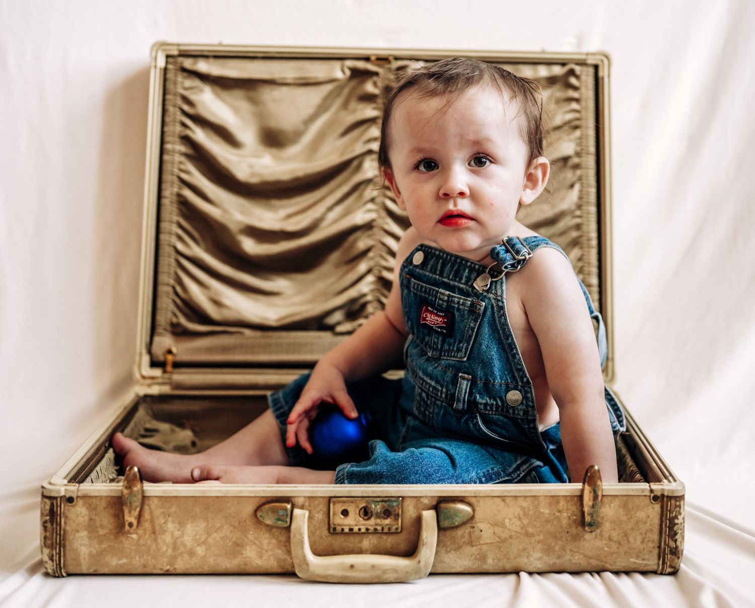 A sequence of four photos showing a baby in a vintage suitcase wearing a teal dress against a cream backdrop.