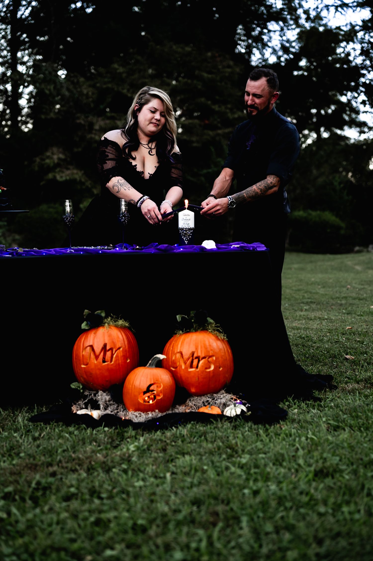 Mr and Mrs carved pumpkins displayed on grass with purple decor and candles.