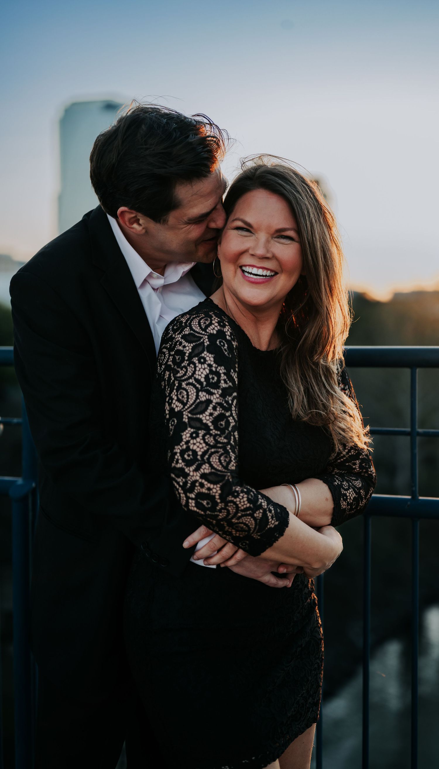 A smiling couple in black outfits share a romantic moment on a rooftop at sunset, man kissing woman's cheek.