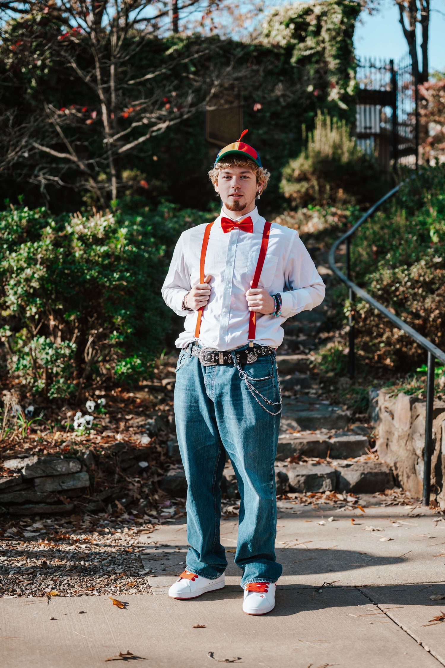 Young man in white shirt, red bow tie, suspenders, feathered cap, and jeans posing outdoors in a park setting.