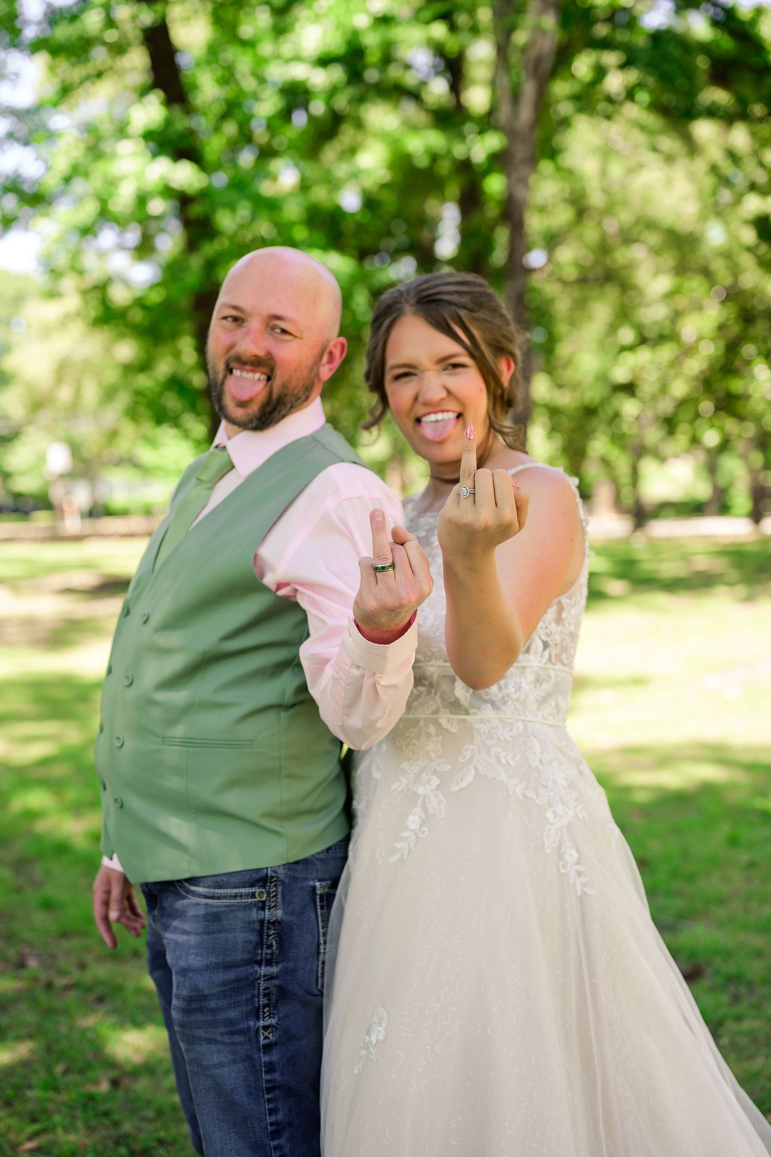 Newlyweds in wedding attire showing off rings while sticking out tongues in a playful outdoor park photo.