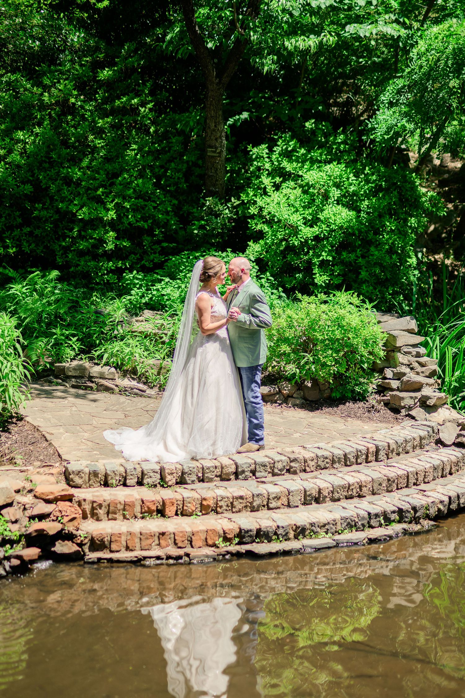 Bride and groom share a romantic moment on a stone platform surrounded by lush greenery and a serene pond.