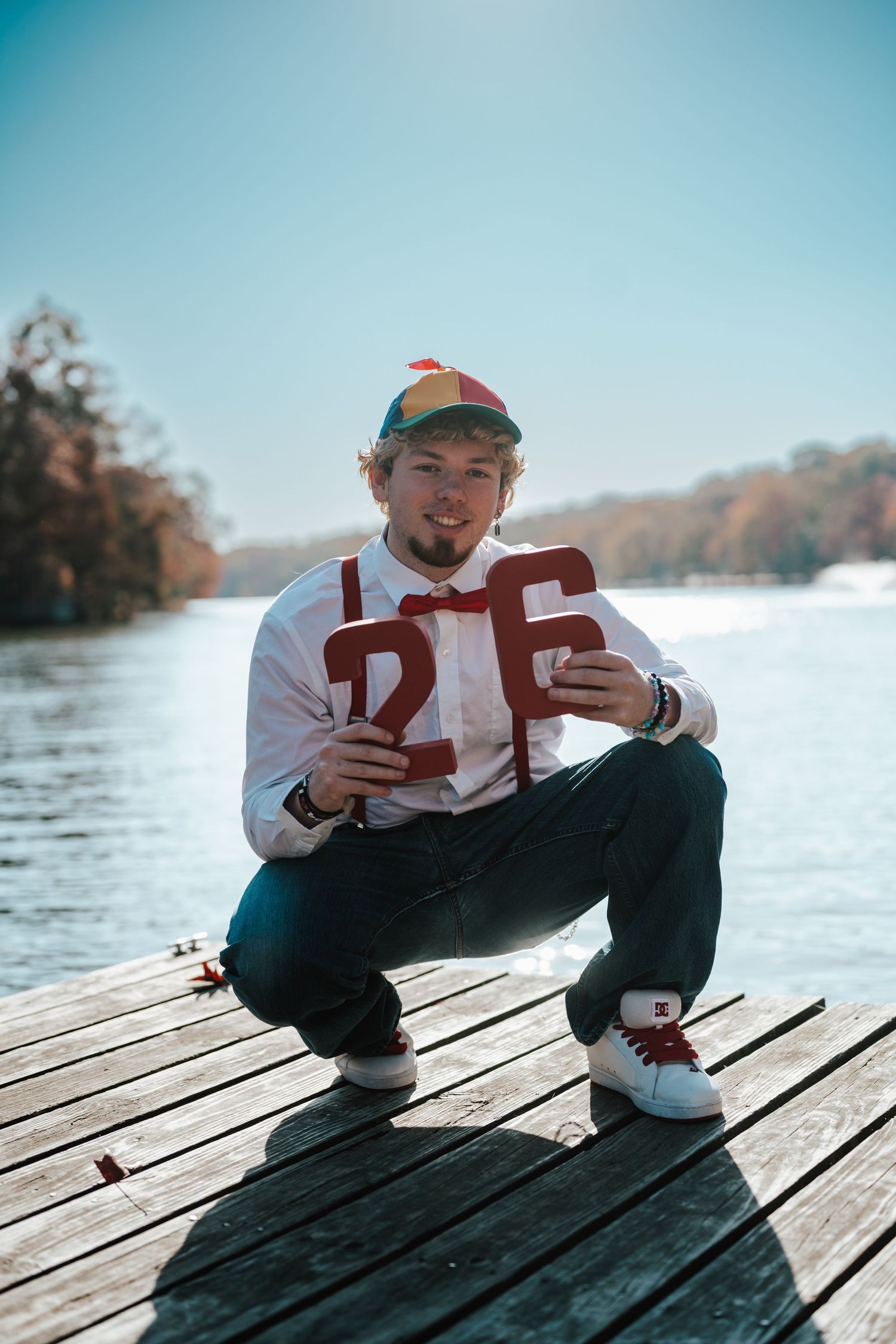 Young man crouching on a lake dock, holding red number 26, wearing a rainbow cap and white shirt on a sunny day.
