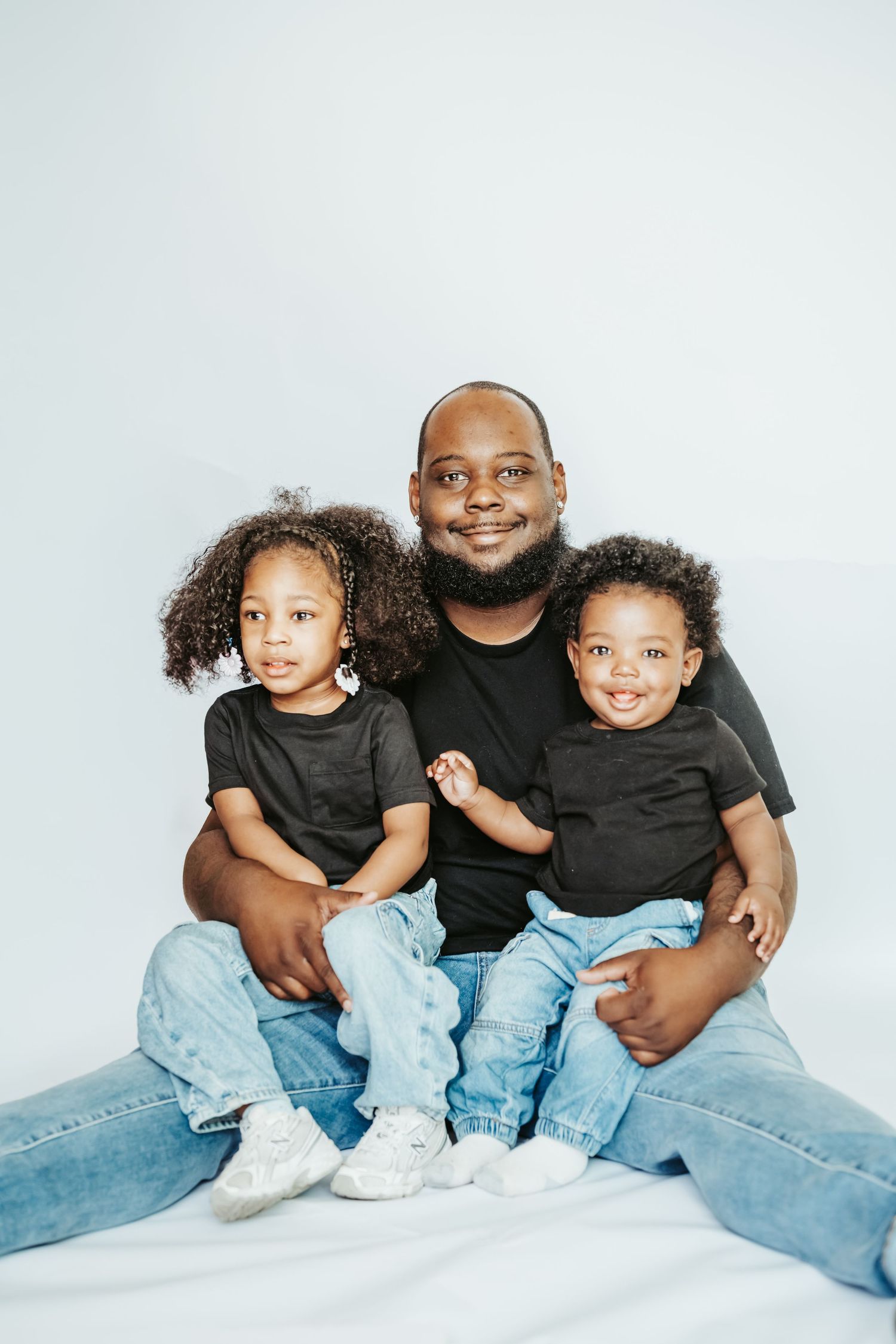 A father sits smiling with his two young children, all wearing black shirts and jeans, against a white background.