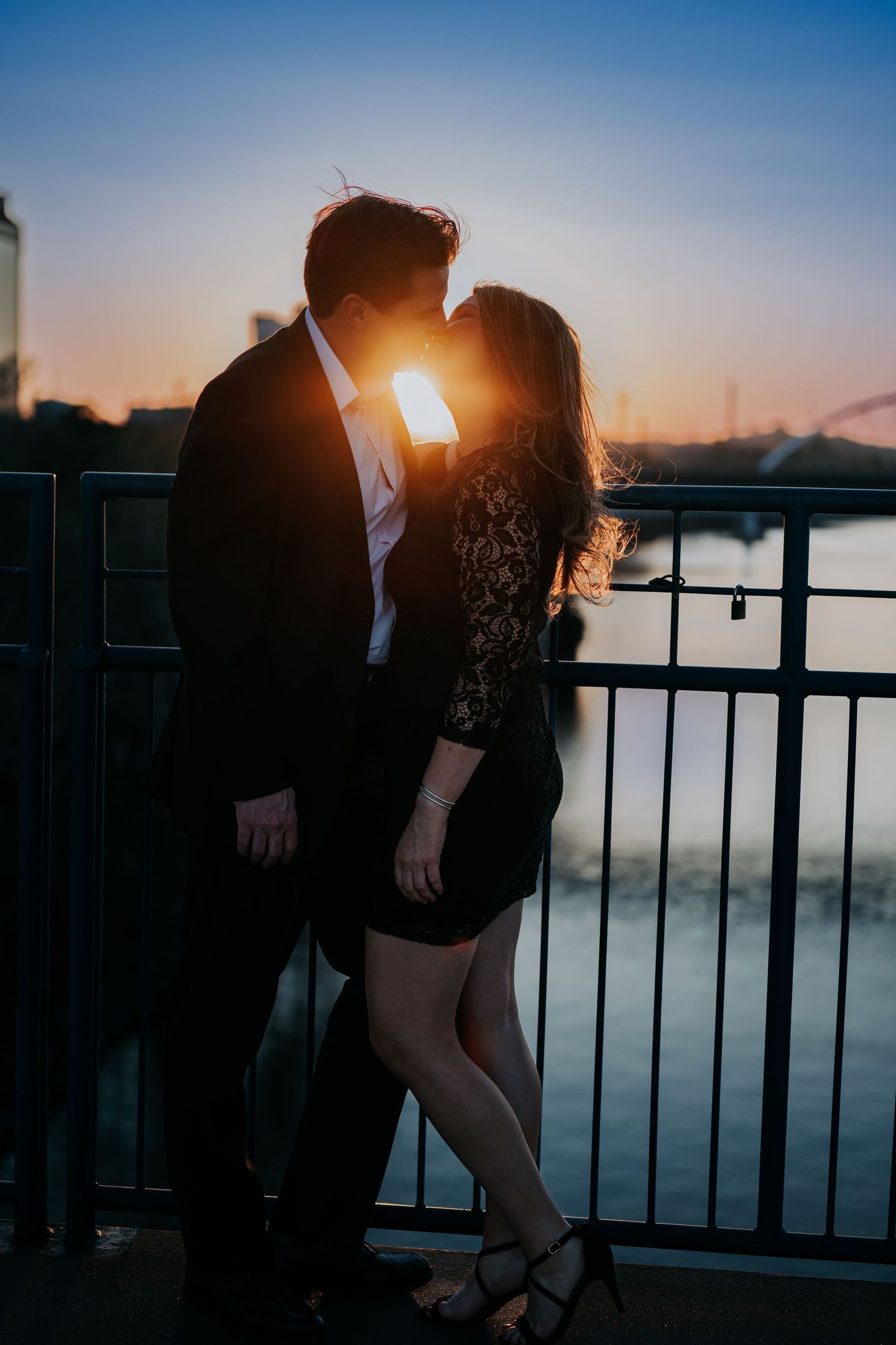 Couple sharing a romantic kiss at sunset on a bridge, silhouetted against a golden sky near a river.