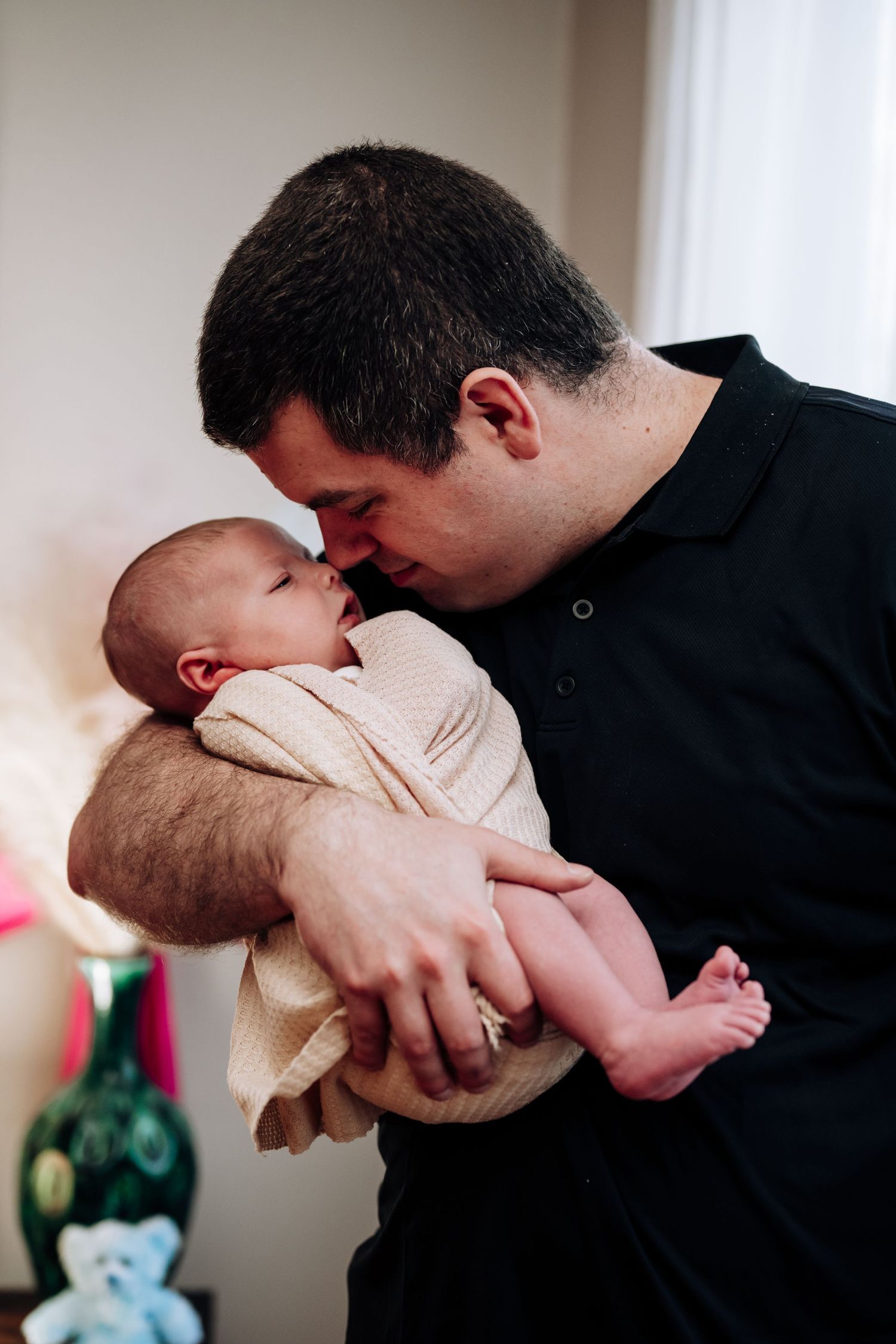 Father in black shirt tenderly touching noses with newborn baby cradled in his arms at home.