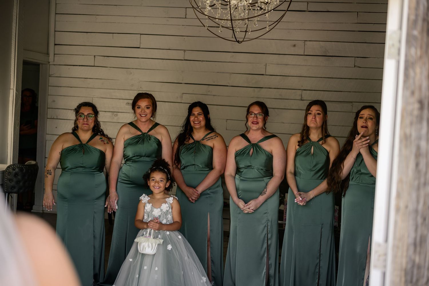 Bridesmaids in sage green dresses react emotionally as flower girl stands in front during wedding ceremony.