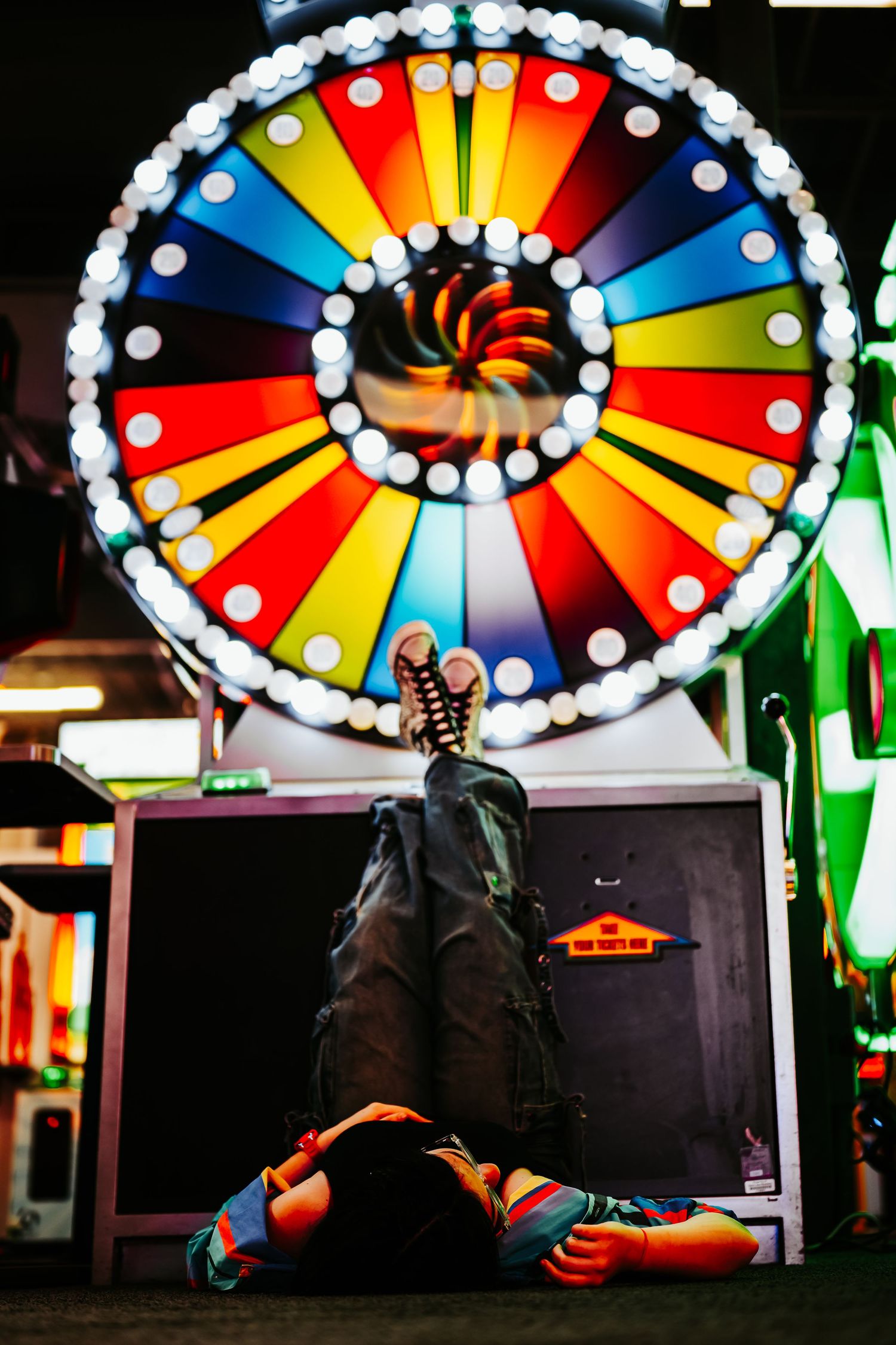 Person lying on floor beneath colorful spinning prize wheel at arcade, feet raised wearing sneakers.