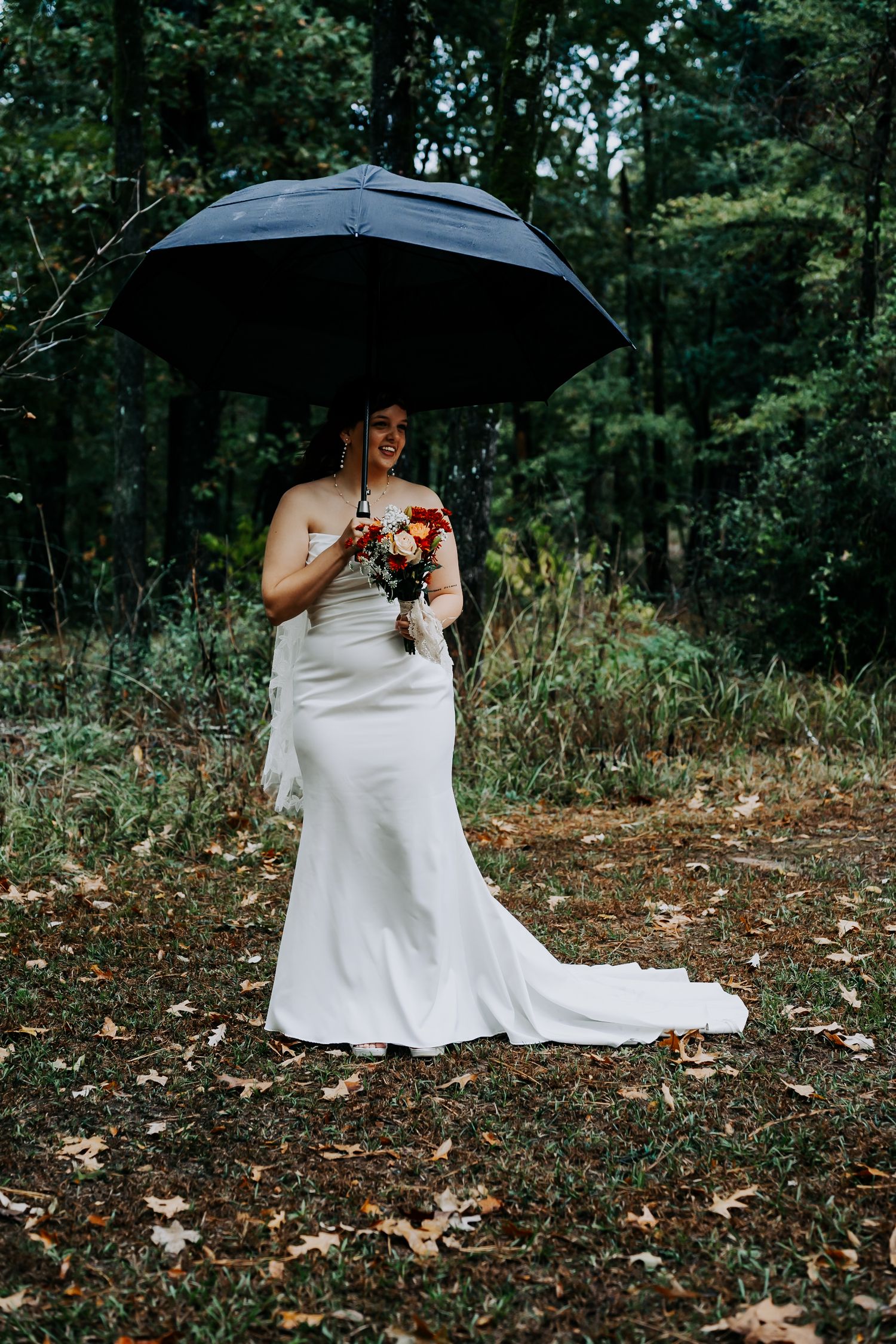 A person in a white wedding dress sits on fallen autumn leaves in a wooded setting holding a black umbrella.