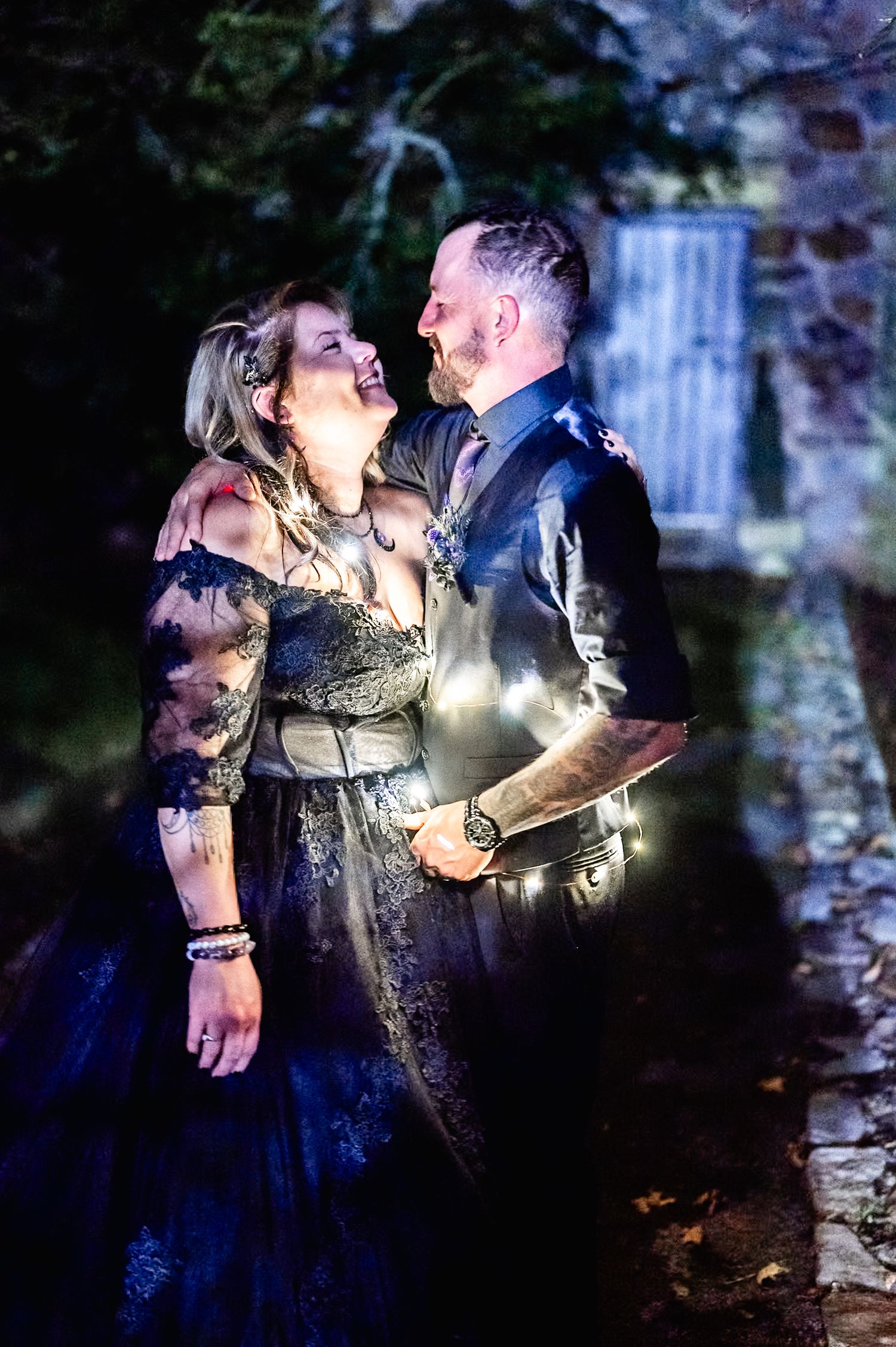 A couple shares a romantic moment in front of a waterfall at night with twinkling lights creating a magical atmosphere.