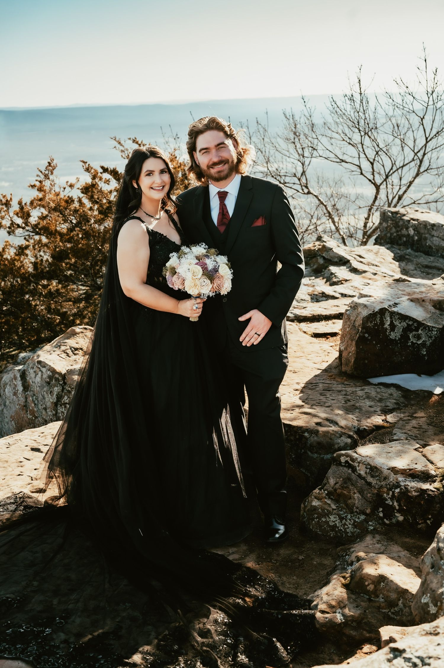 Couple in formal wear posing together on rocky mountain ledge during sunset.
