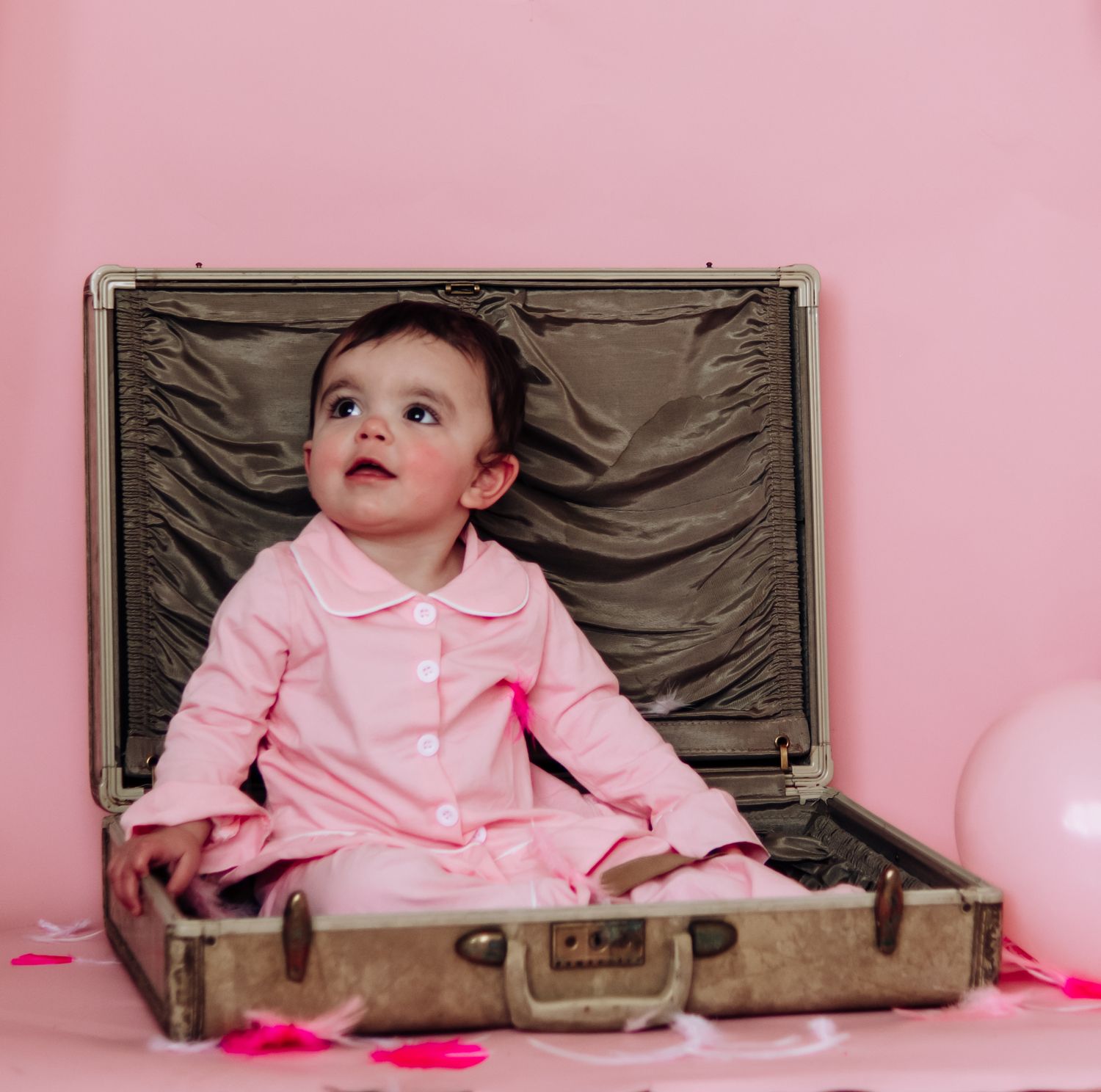 Baby wearing pink outfit sits in vintage brown suitcase against pink background in progressive sequence.