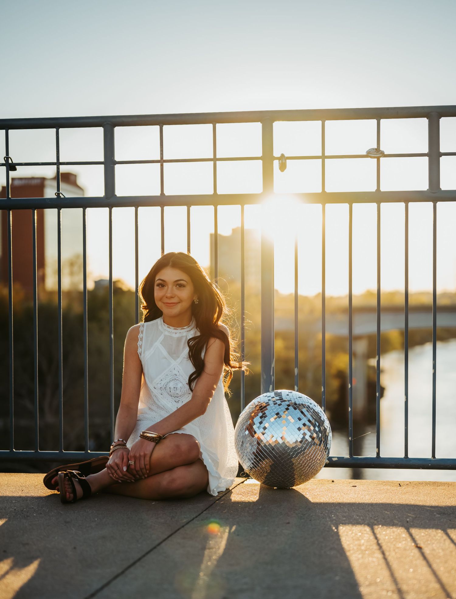 Young woman in white dress sitting by iron railing at sunset with a silver disco ball beside her.