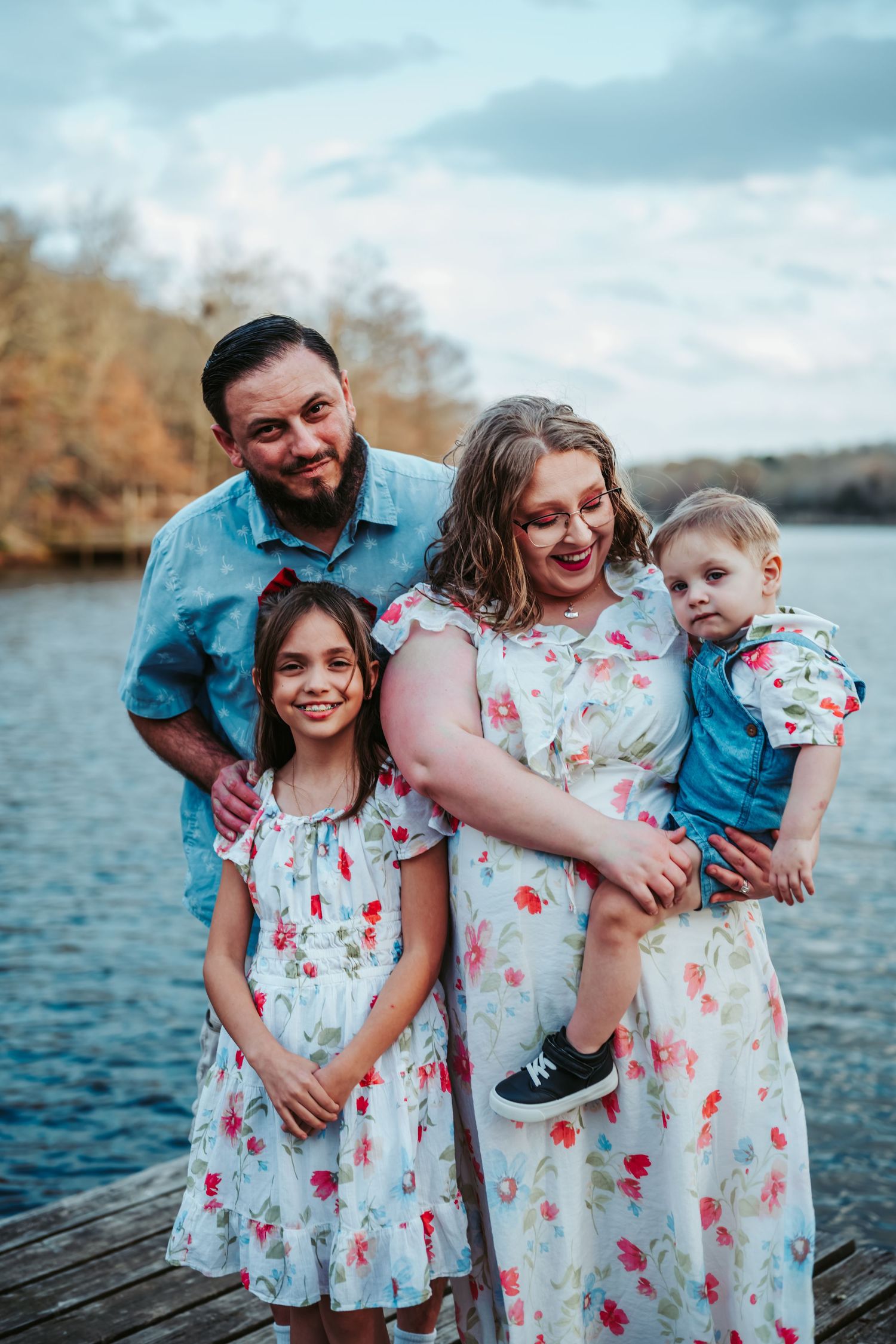 Happy family of four posing on wooden dock by lake in coordinating floral outfits.