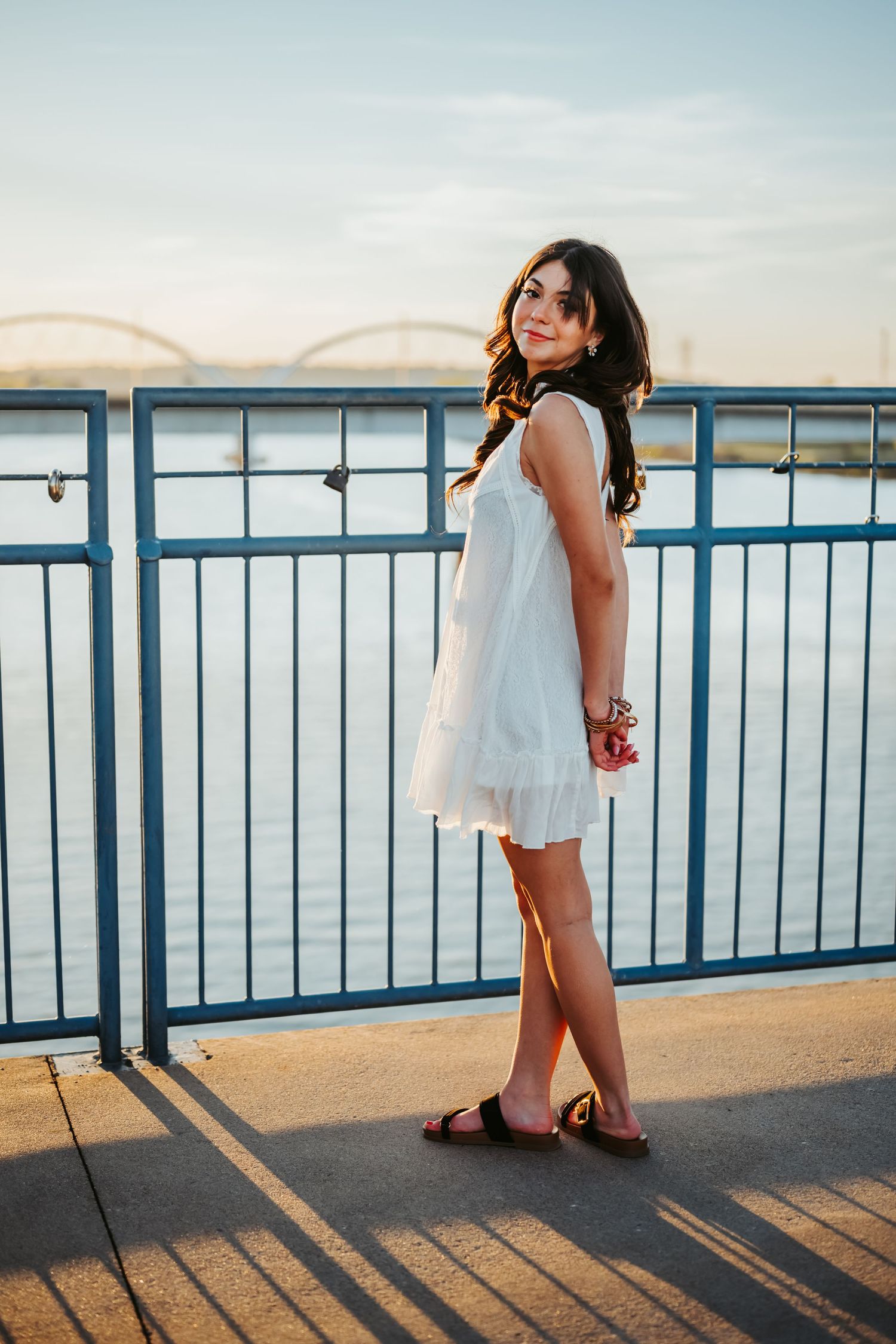Young woman in white dress posing on a bridge railing with a river and arch bridge at sunset.