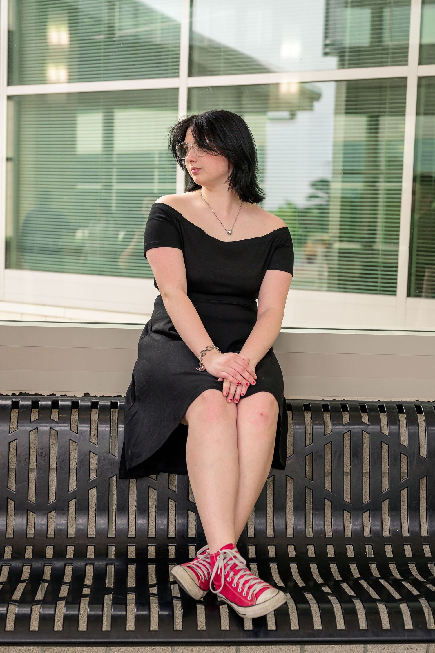 Young woman in black off-shoulder dress and pink sneakers sitting on bench outdoors.