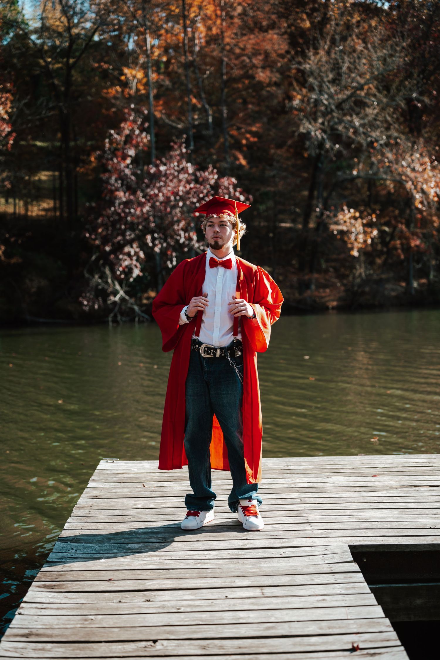 Young man in red graduation gown and cap standing on wooden dock by lake surrounded by autumn foliage.