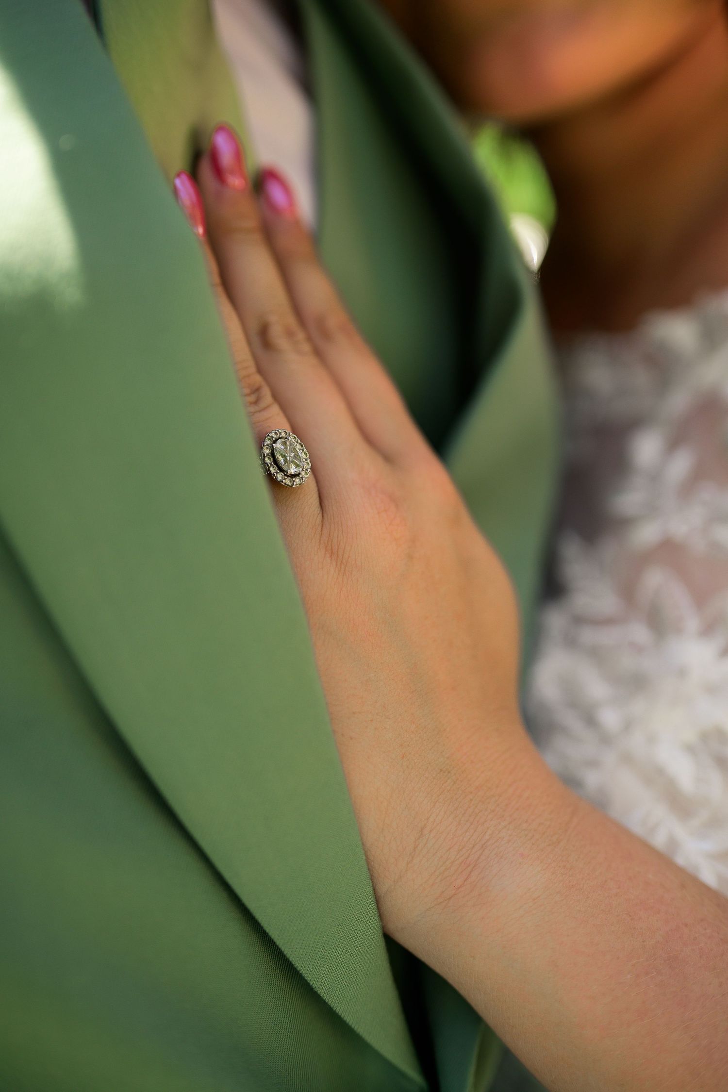 Bride displaying elegant diamond halo engagement ring with pink nails against tropical green leaves.