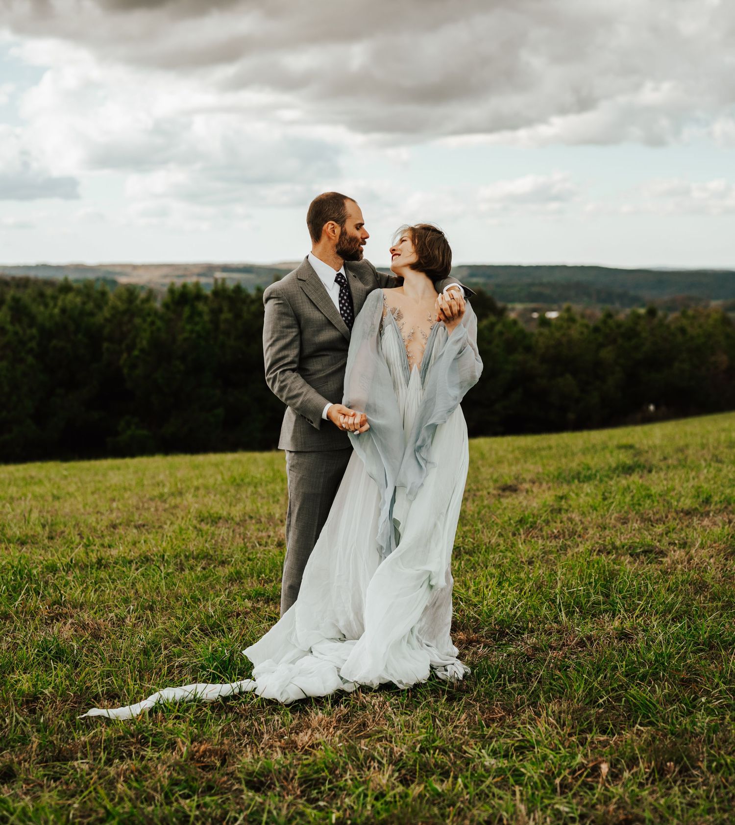 A bride and groom shares an intimate moment on a hilltop overlooking a scenic forest landscape on their wedding day.