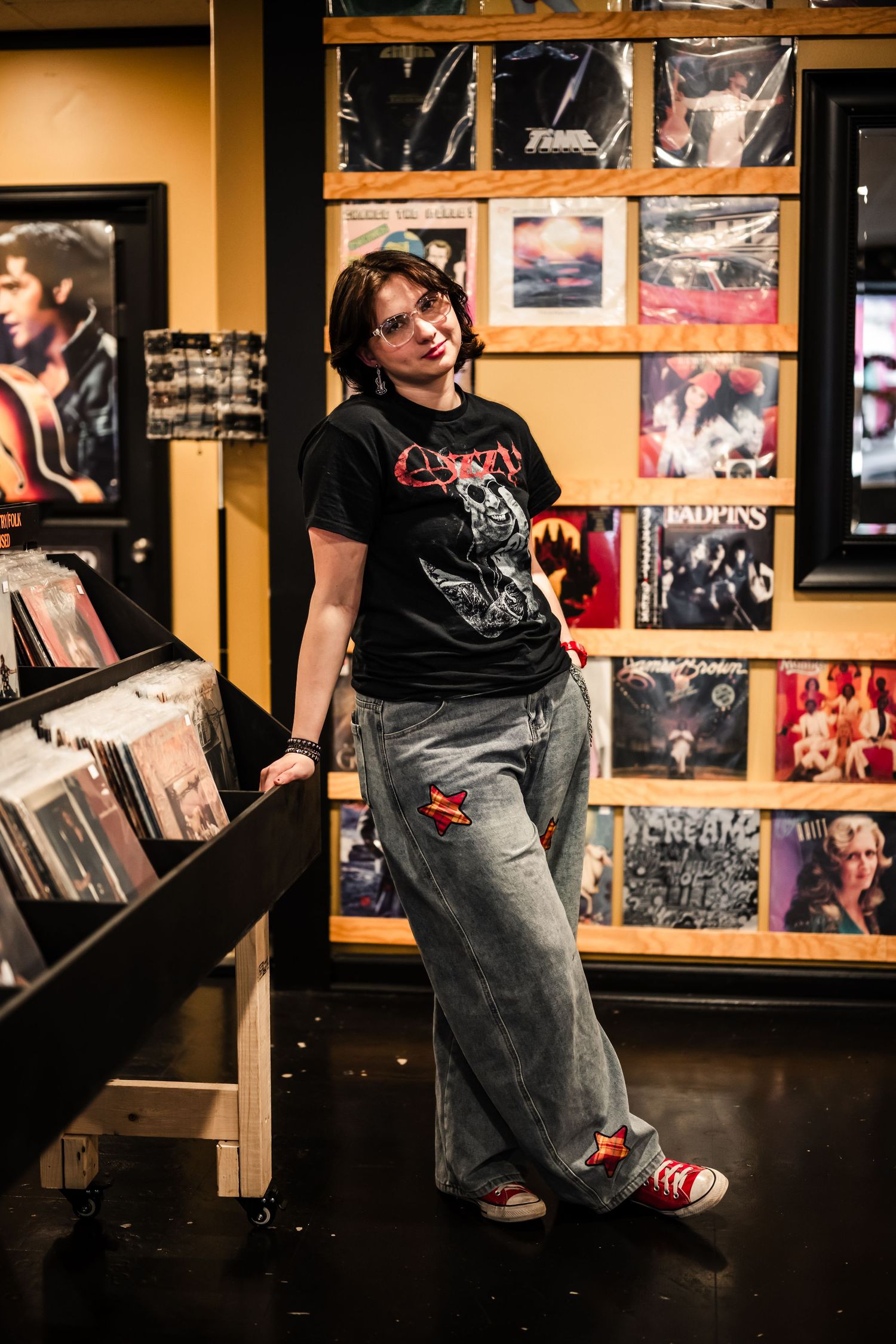 Young woman in band tee and wide-leg jeans poses in a vinyl record store surrounded by album covers.
