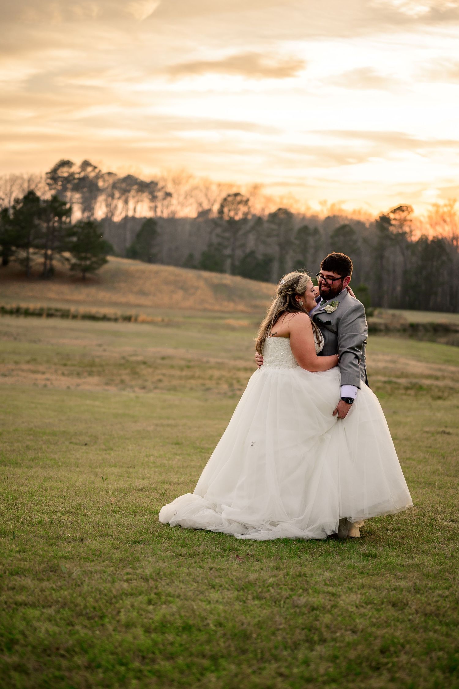 Bride and groom share a romantic embrace at sunset in a green field, with trees and golden sky in the background.