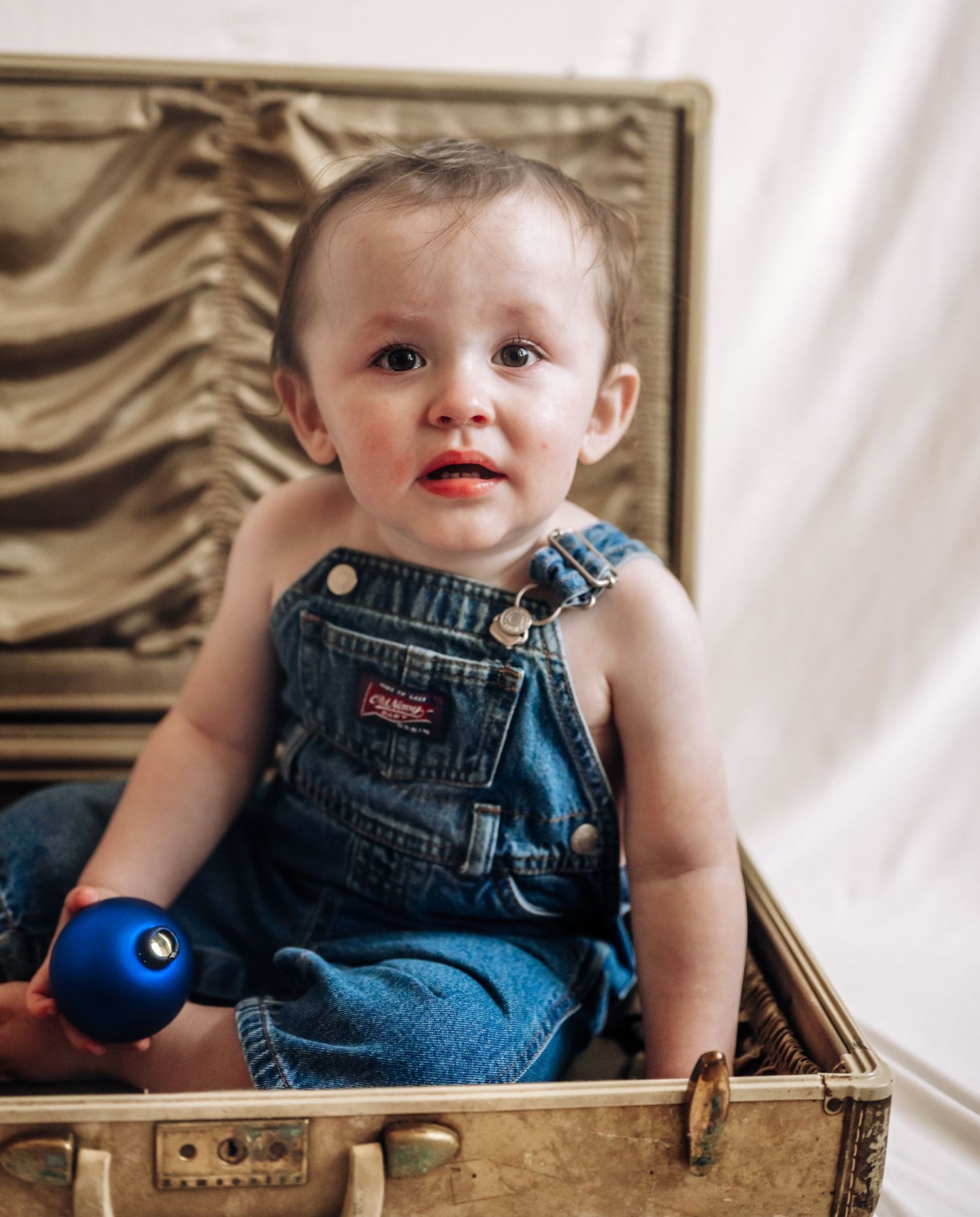 Baby wearing denim overalls sits in a wooden crate with a blue ball toy against a white backdrop.