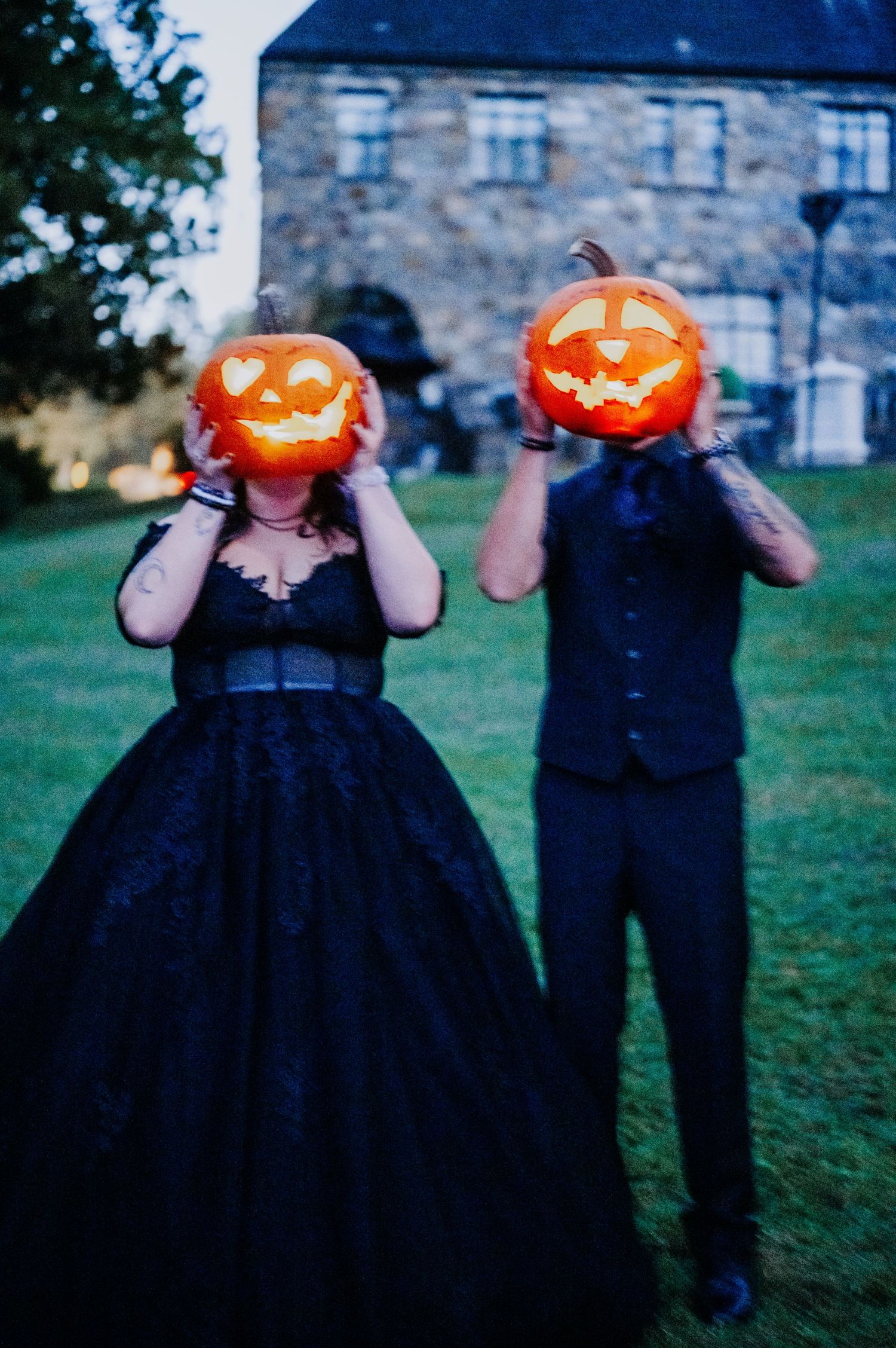 a couple at their wedding, pose outdoors holding illuminated jack-o'-lantern pumpkins in place of their heads.