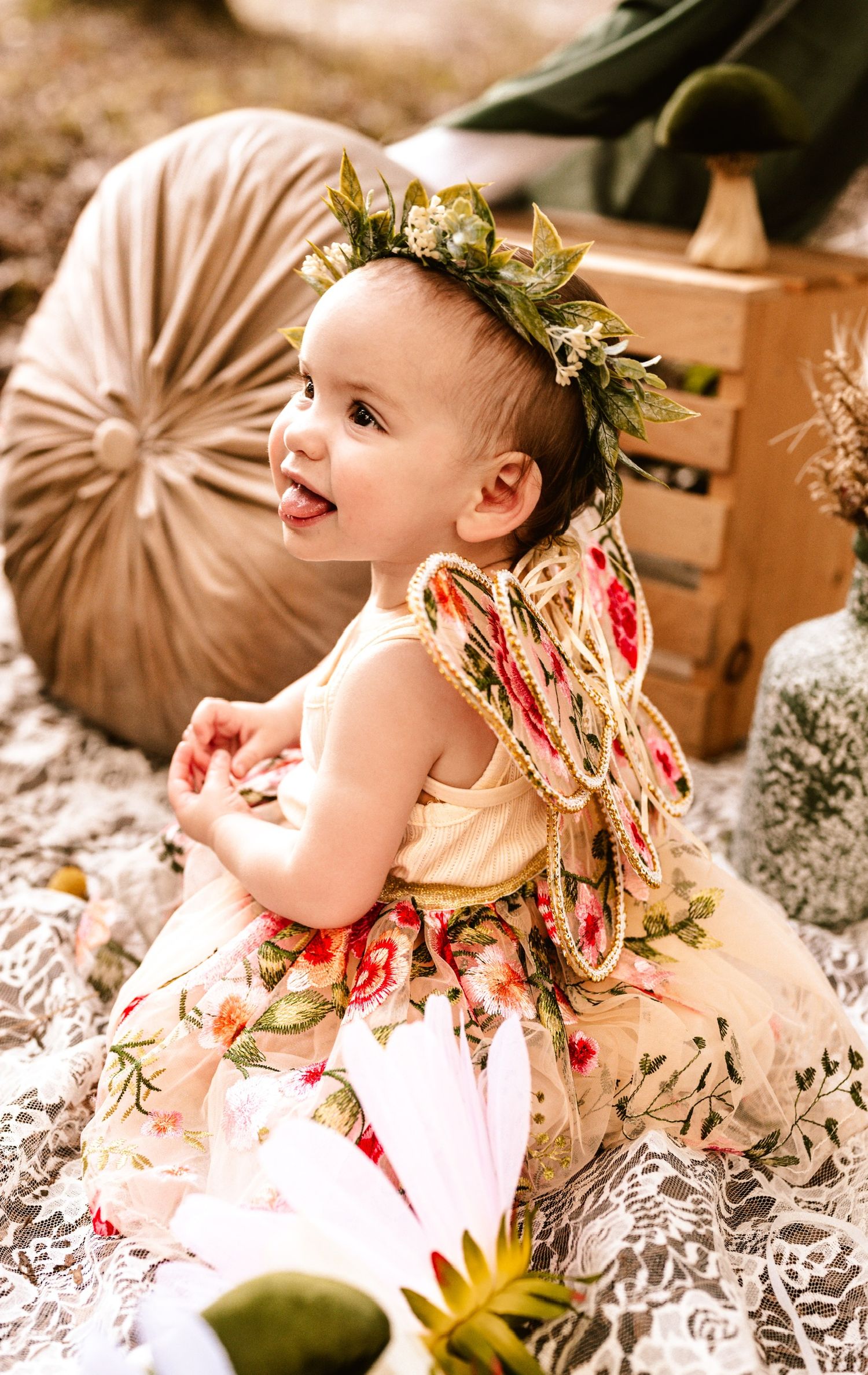 Baby wearing floral crown and dress smiles while lying on textured blanket.