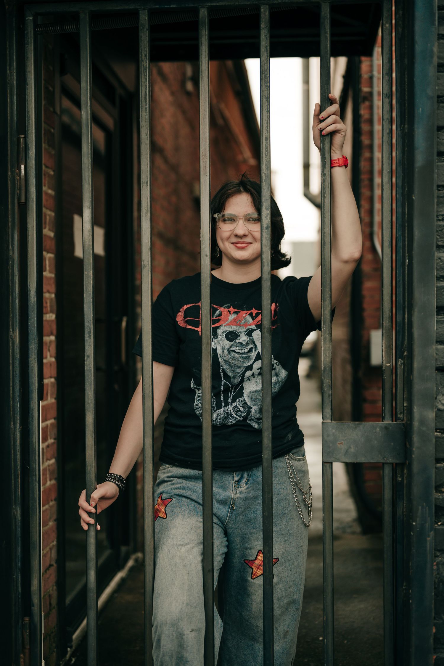 Young woman in black band tee and jeans posing between metal bars in a brick alley.