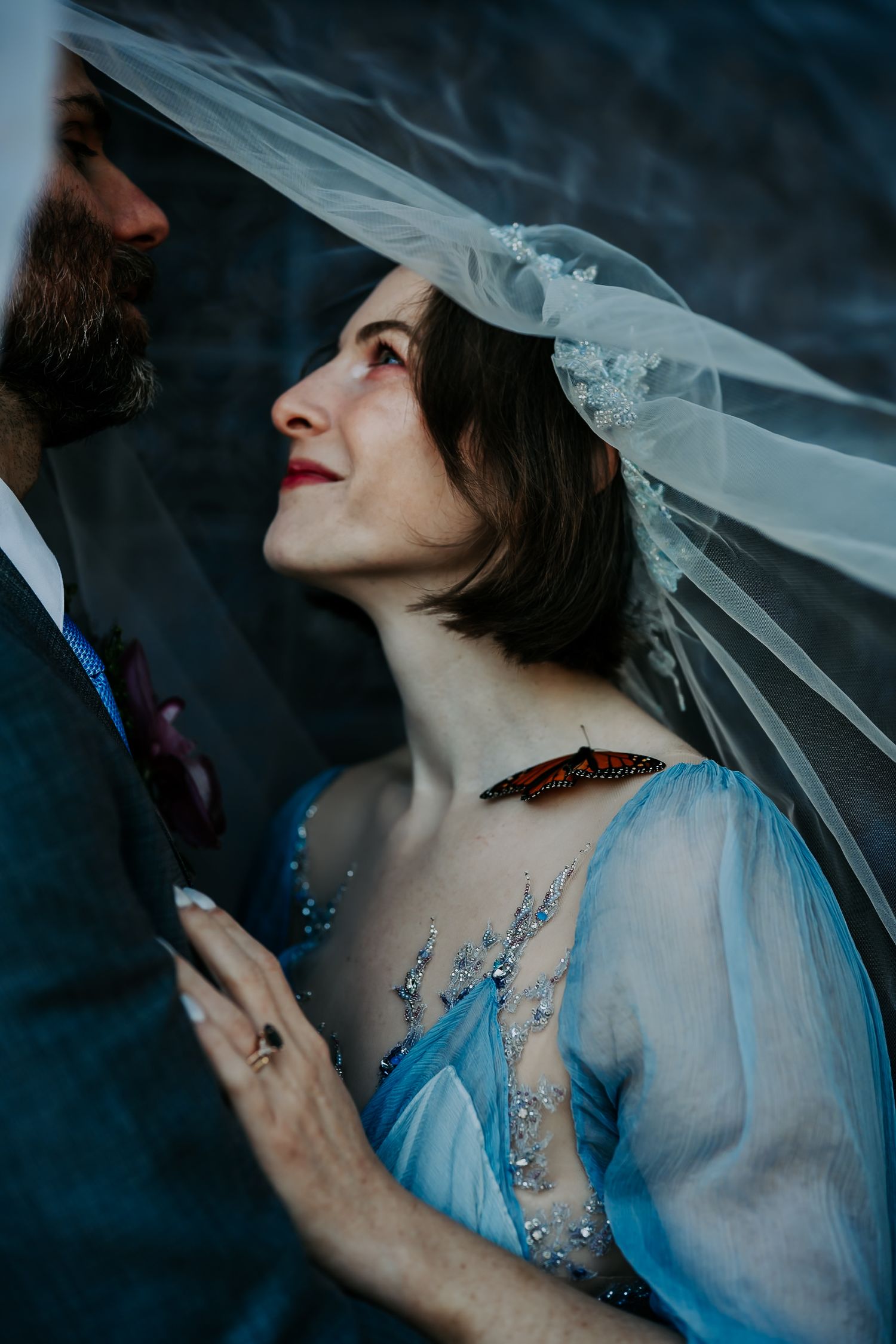 A romantic wedding moment captured in moody lighting shows a bride in a blue dress and veil looking up lovingly.