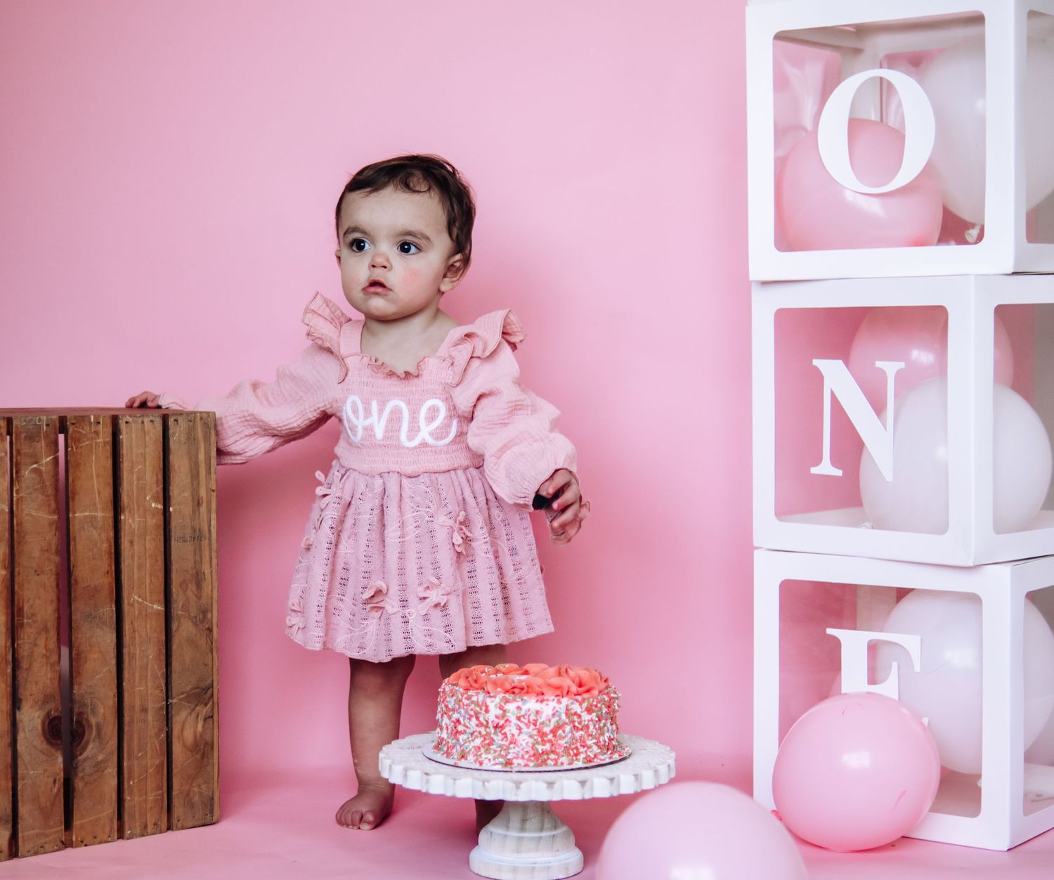 Pink first birthday party setup with decorative letter blocks, wooden crate, and pink smash cake on white pedestal.