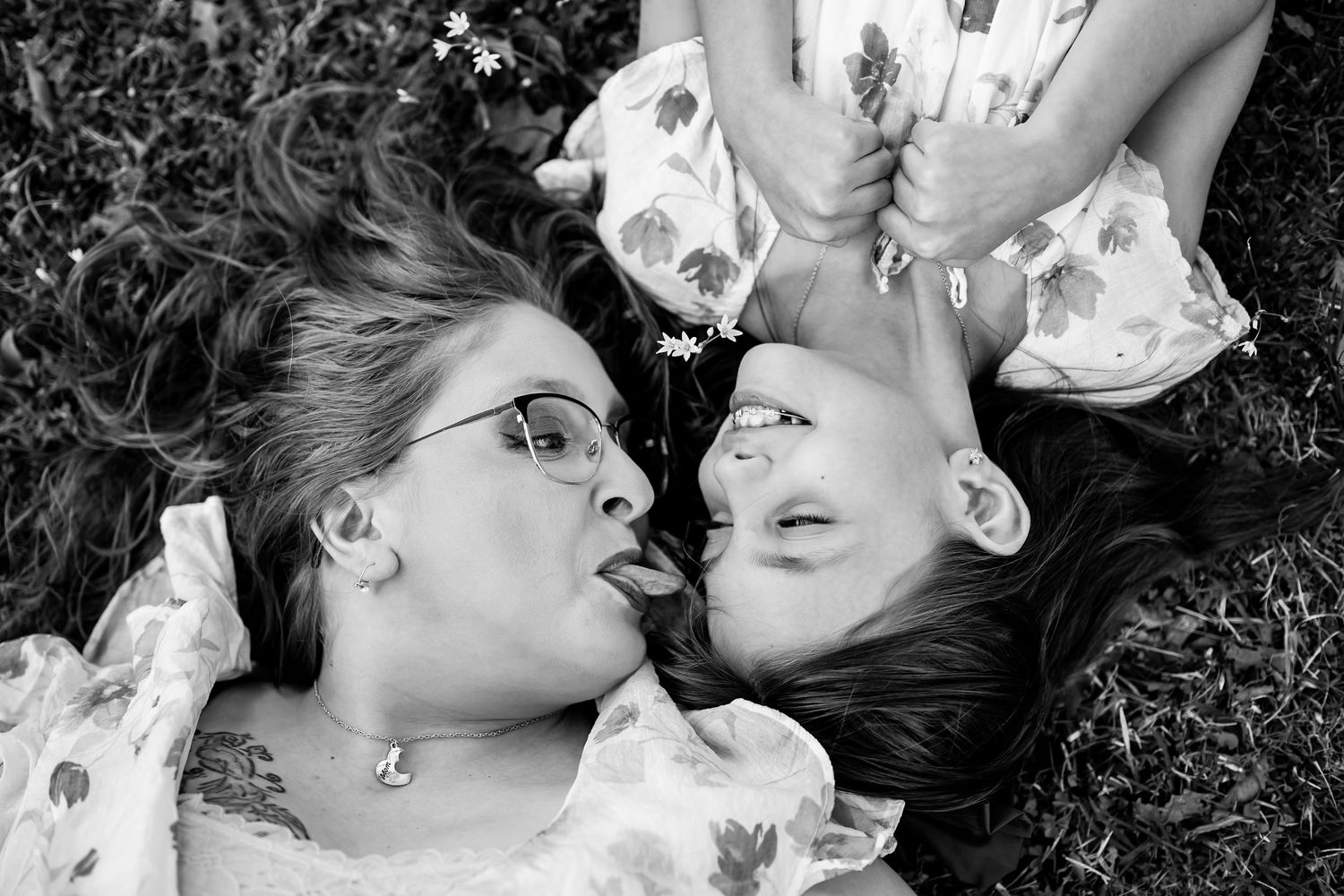 Black and white photo of two women lying on grass, laughing and sticking out tongues, wearing floral dresses.