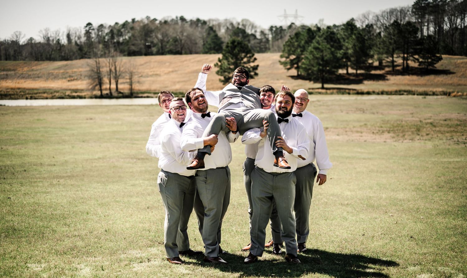 Groomsmen lifting groom in gray suits with bow ties on green field at outdoor wedding celebration.