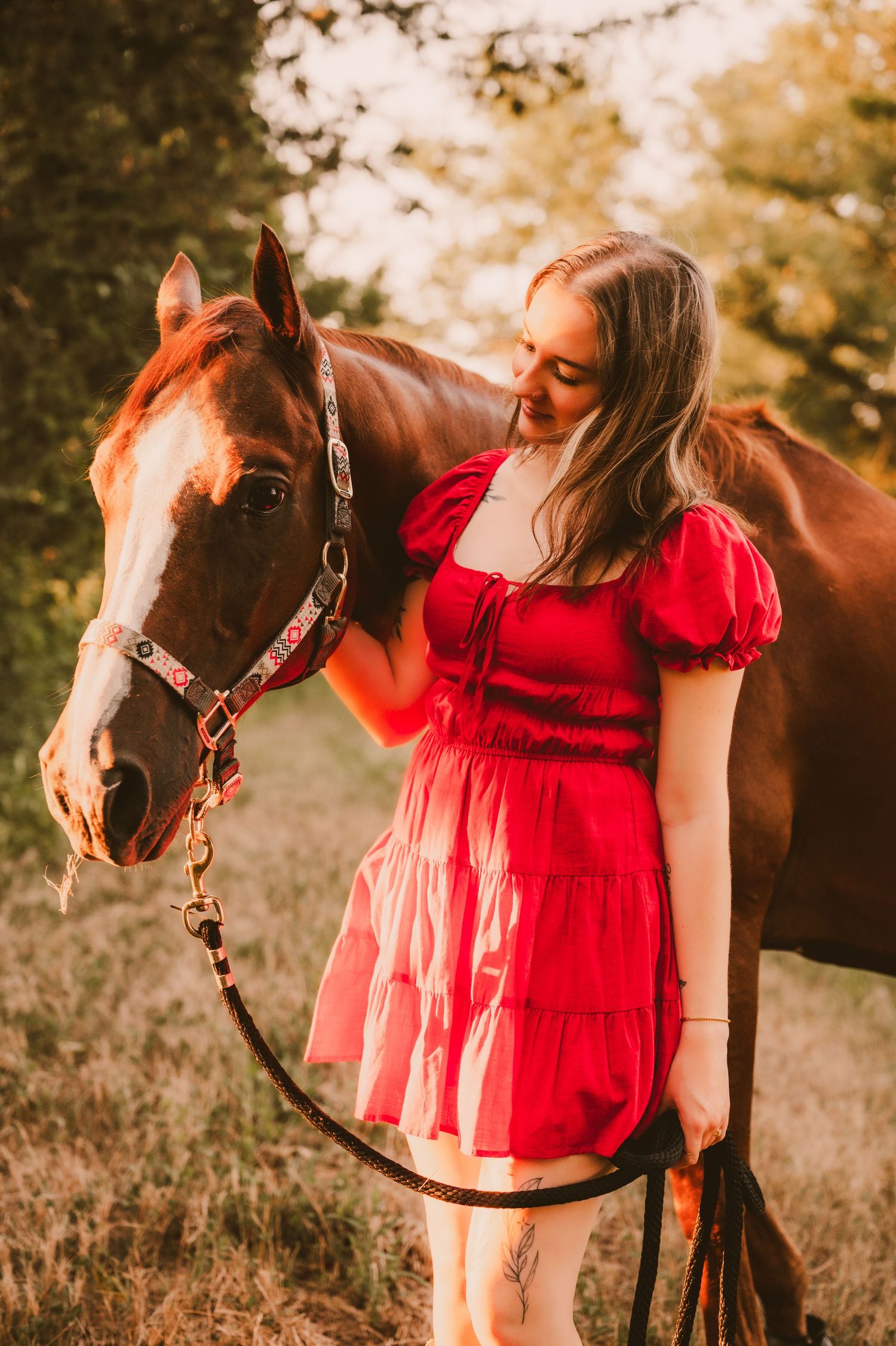 Woman in red dress poses with brown horse during golden hour photoshoot.