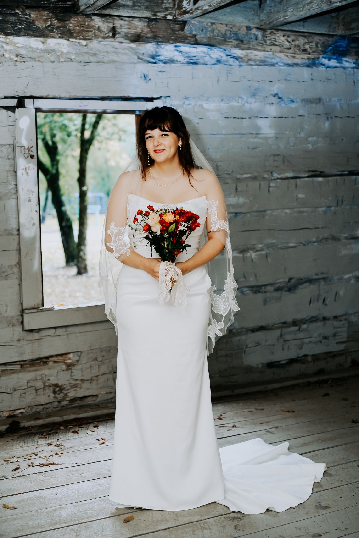 Bride in white lace gown sitting in rustic wooden building with natural light.