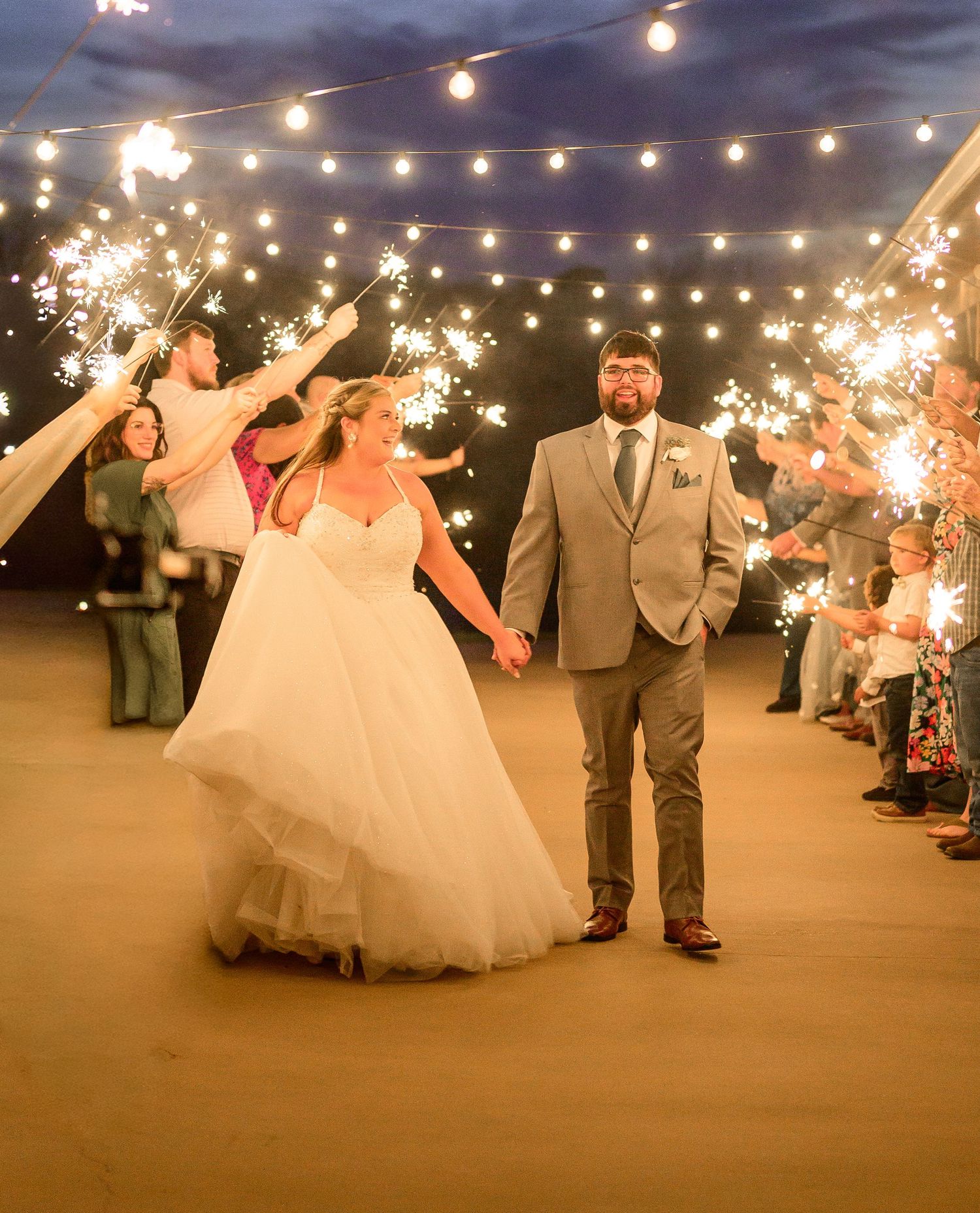 Bride and groom walk through sparkler exit under string lights at outdoor evening wedding celebration.